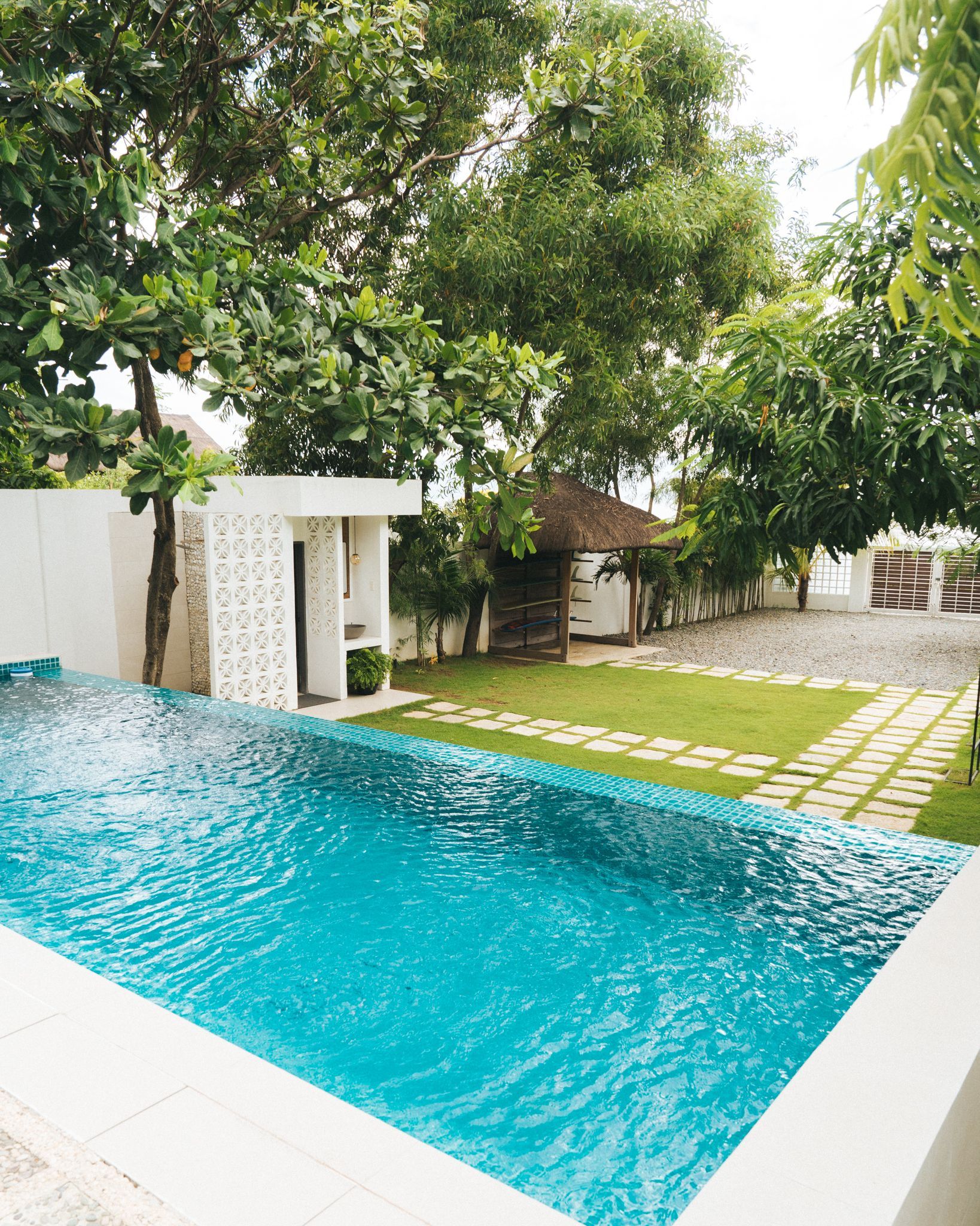 Swimming pool next to a white wall with lush green foliage, leading to a small building and yard.