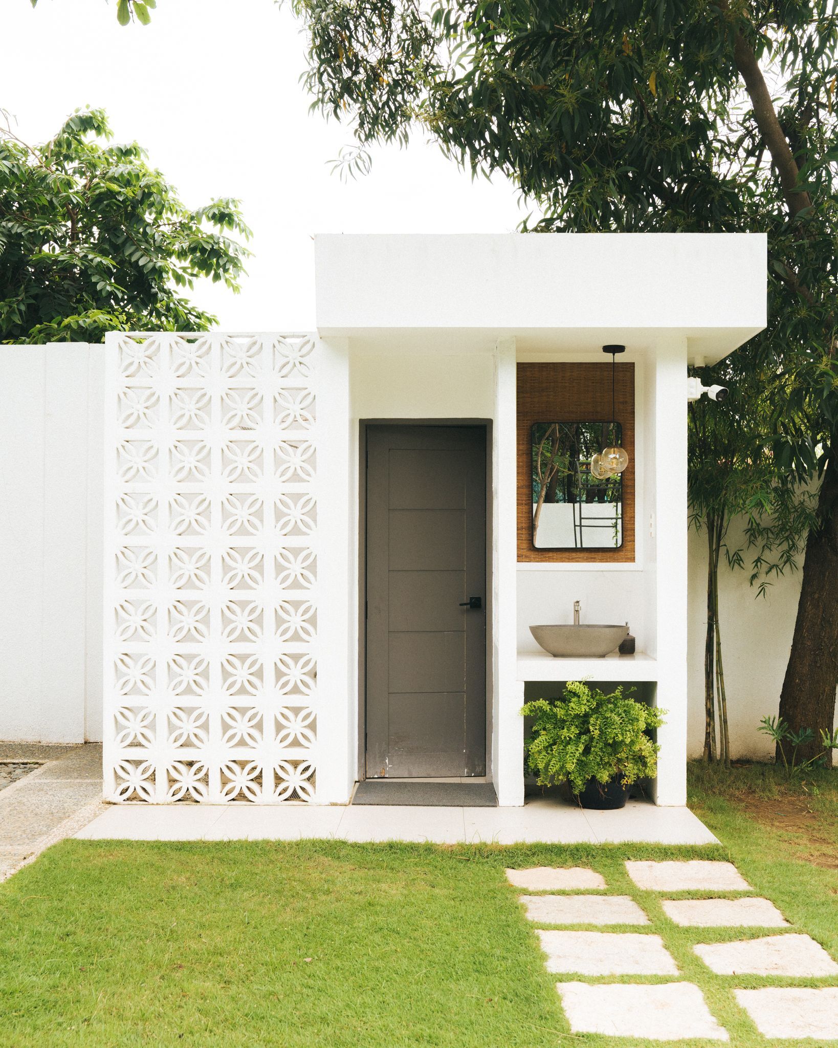 Small white outdoor structure with gray door, open sink area, and patterned wall, set on a green lawn.