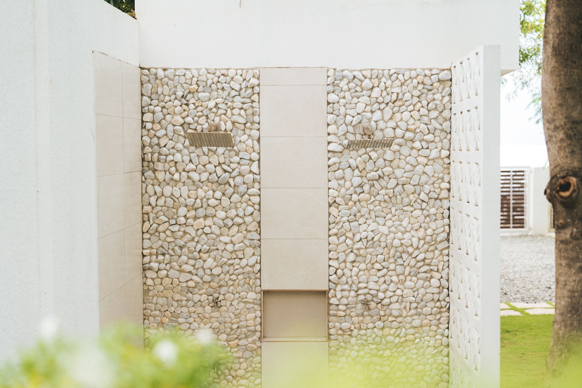 White outdoor shower with stone walls, a central panel, and a glimpse of greenery.