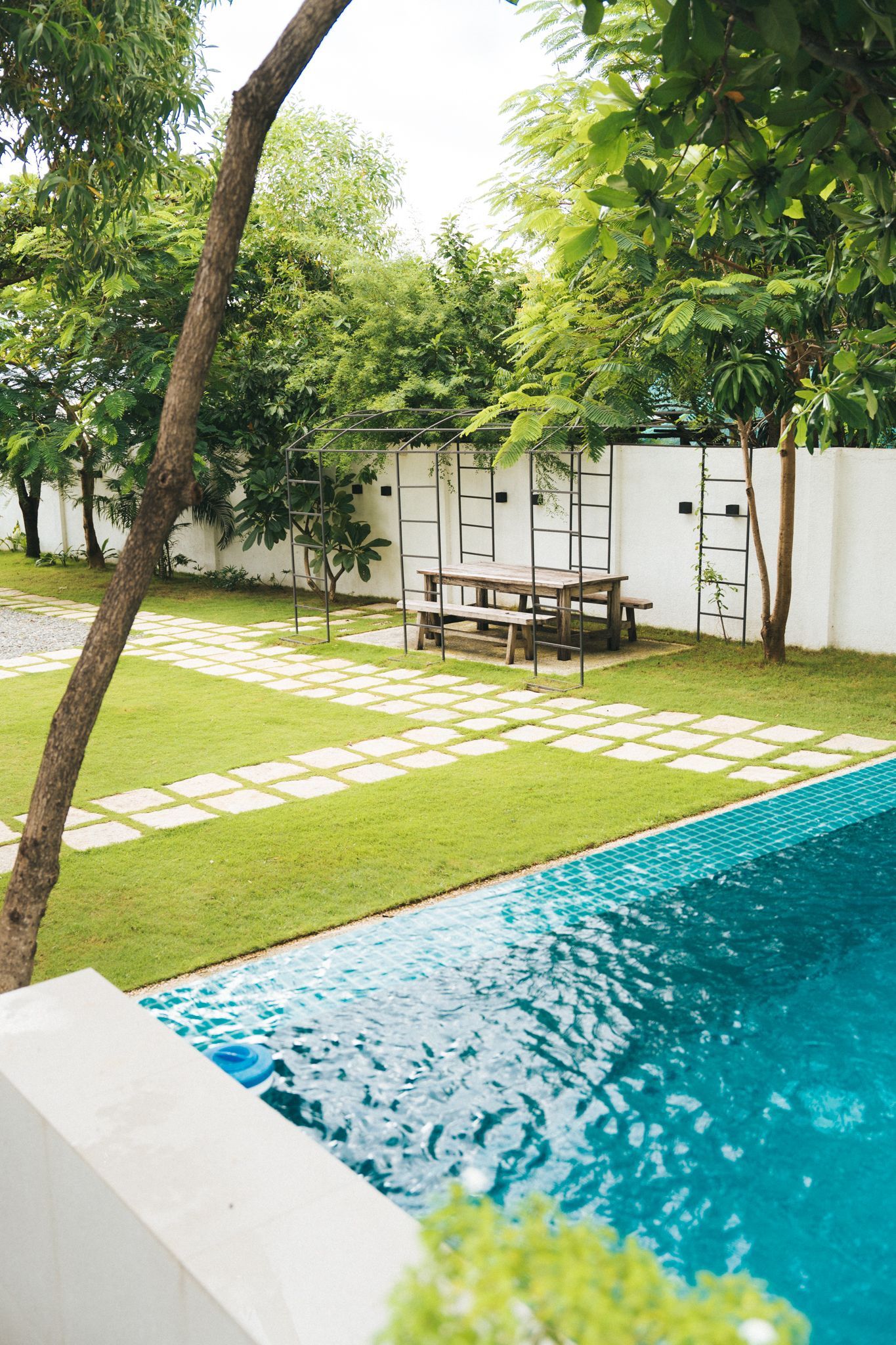 Lush backyard with a pool, grassy areas, stone pathways, a wooden picnic table, and surrounding trees.