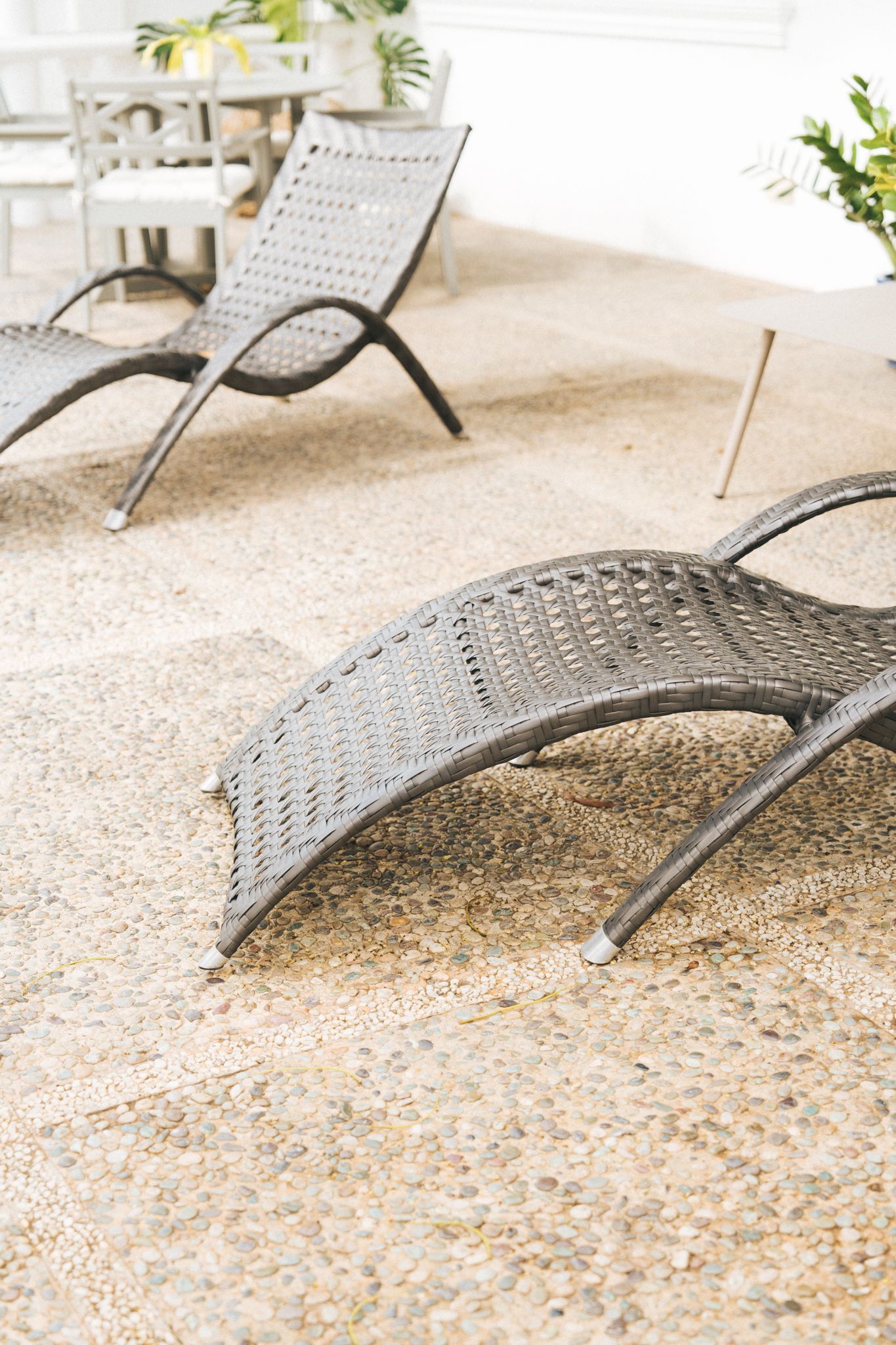 Two dark wicker chaise lounge chairs on a gravel patio, with other outdoor furniture and plants in the background.