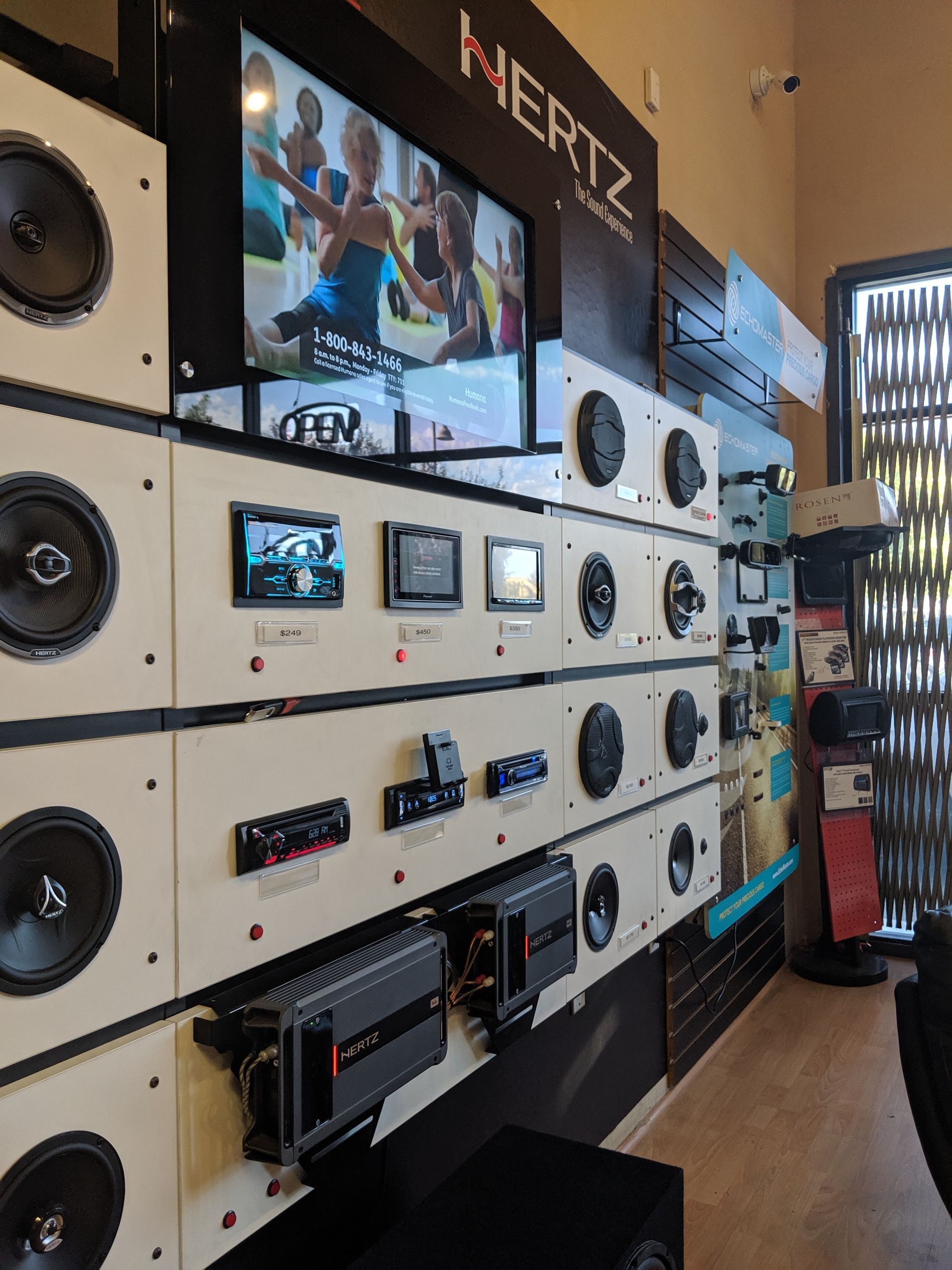 Wall-mounted display of audio equipment and speakers in a showroom, with a TV screen above.