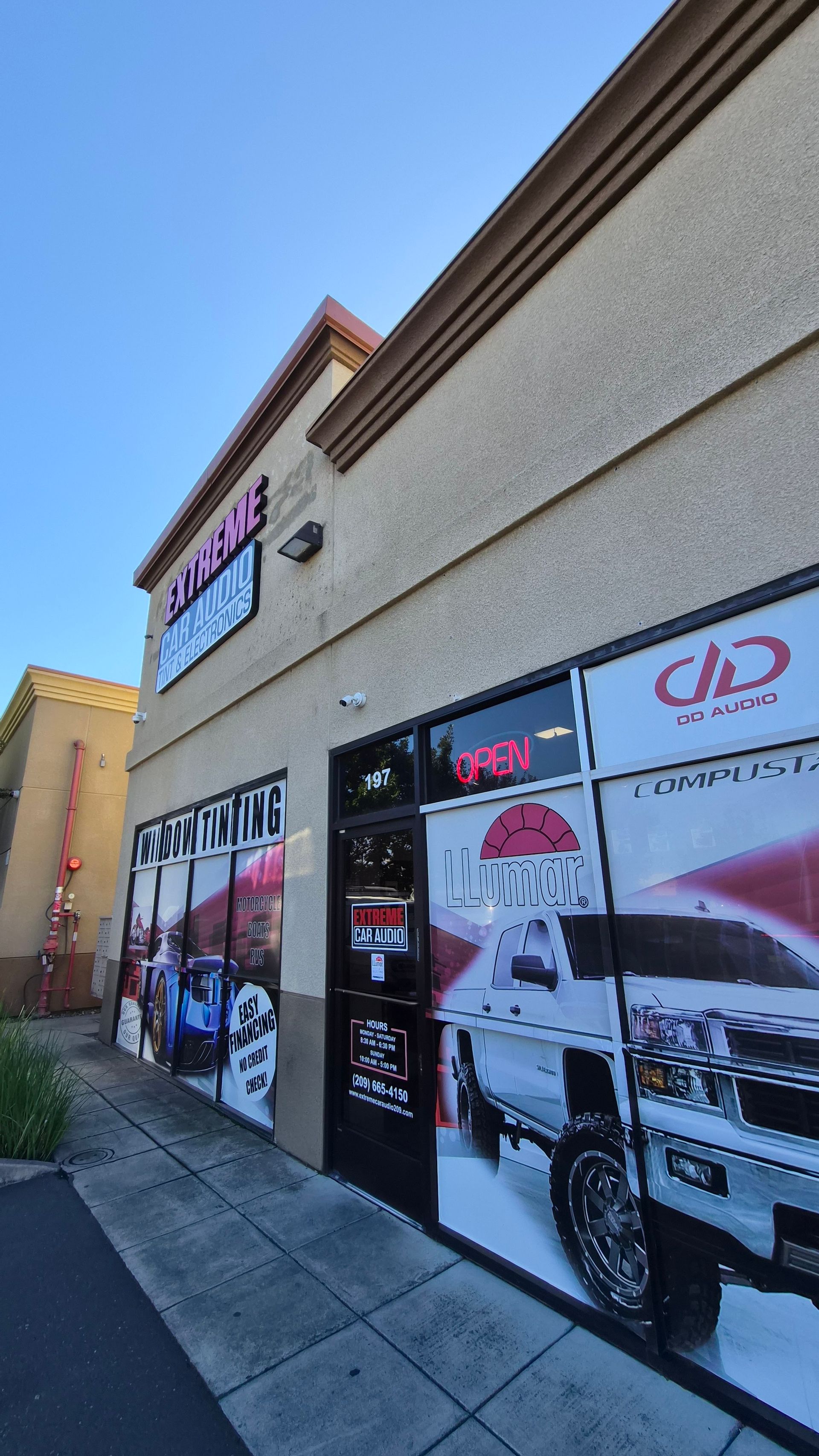 Strip mall storefront with car graphics on windows and a sidewalk outside.