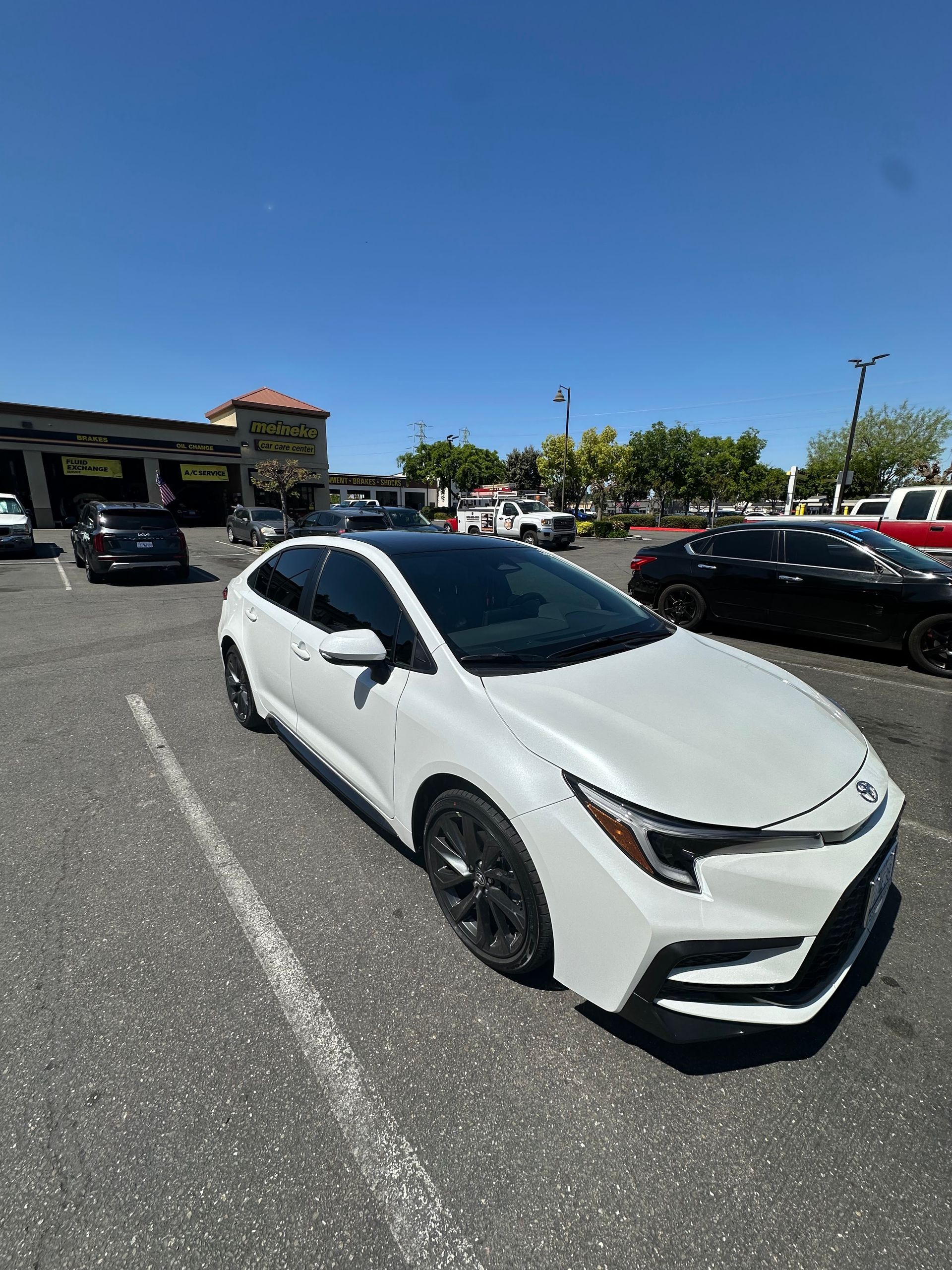 White compact car parked in a sunny parking lot with other vehicles and a clear blue sky