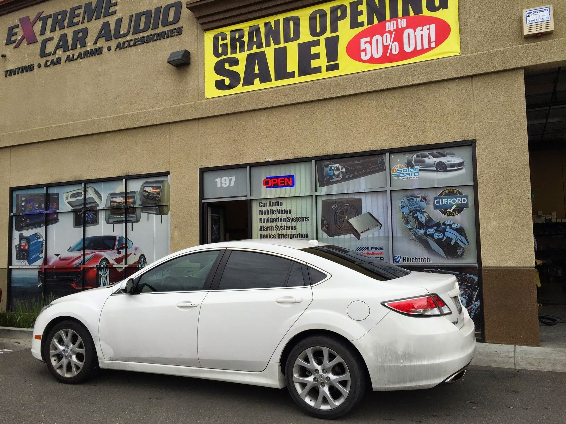White sedan parked outside Extreme Car Audio store with a grand opening sale banner