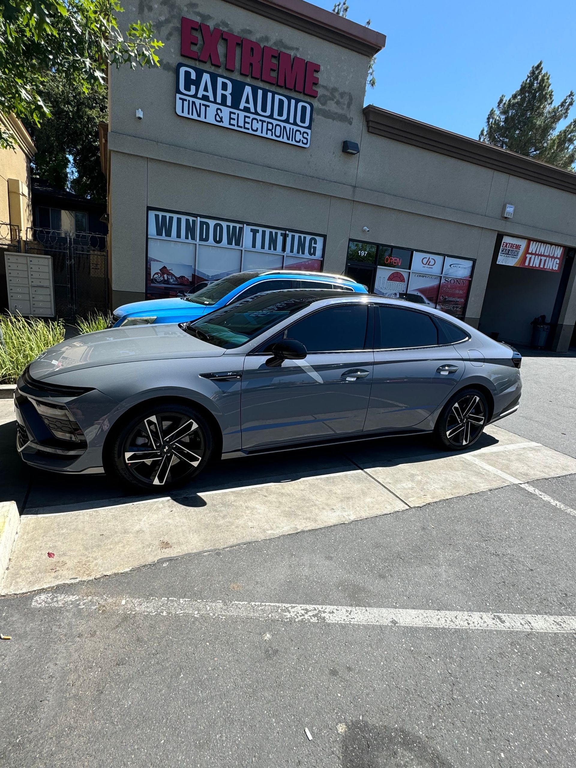 Gray sedan parked outside a car audio shop on a sunny day.