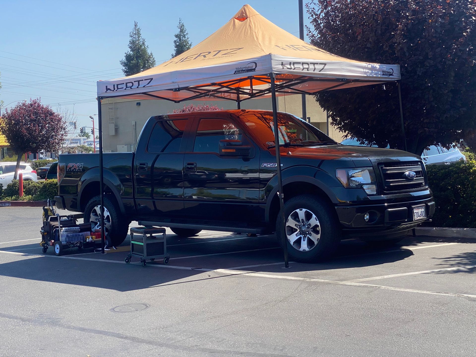 Black pickup truck under an orange canopy tent in a parking lot, with tools and gear nearby