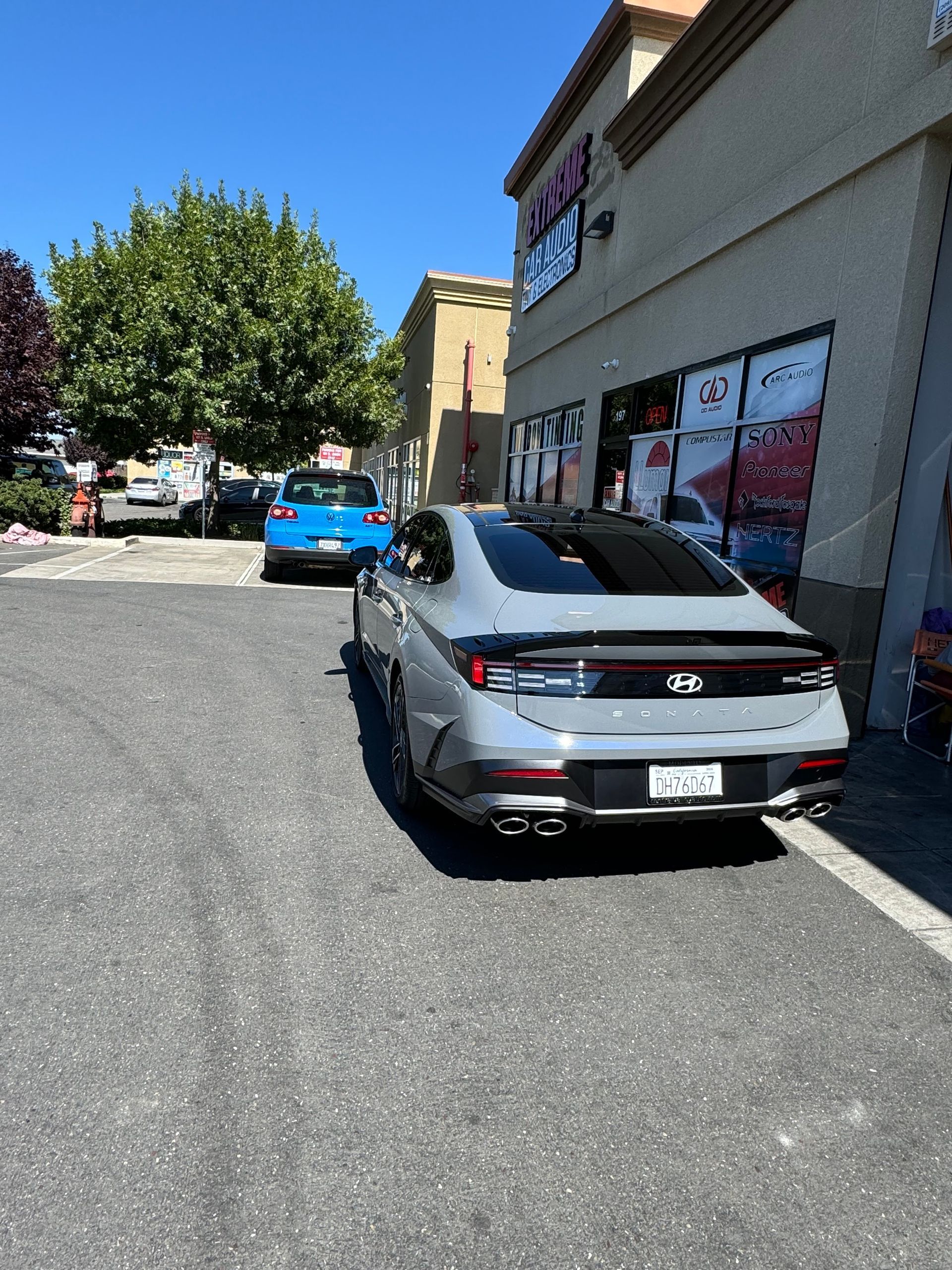 Silver sports car parked beside a storefront on a sunny street, with a blue car ahead.