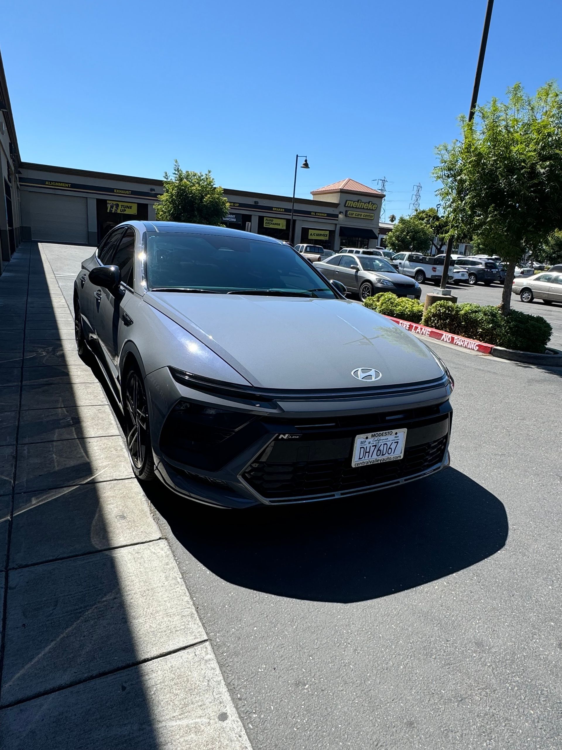 Gray car parked on a steep curbside driveway in bright sunlight, with buildings and trees in the background