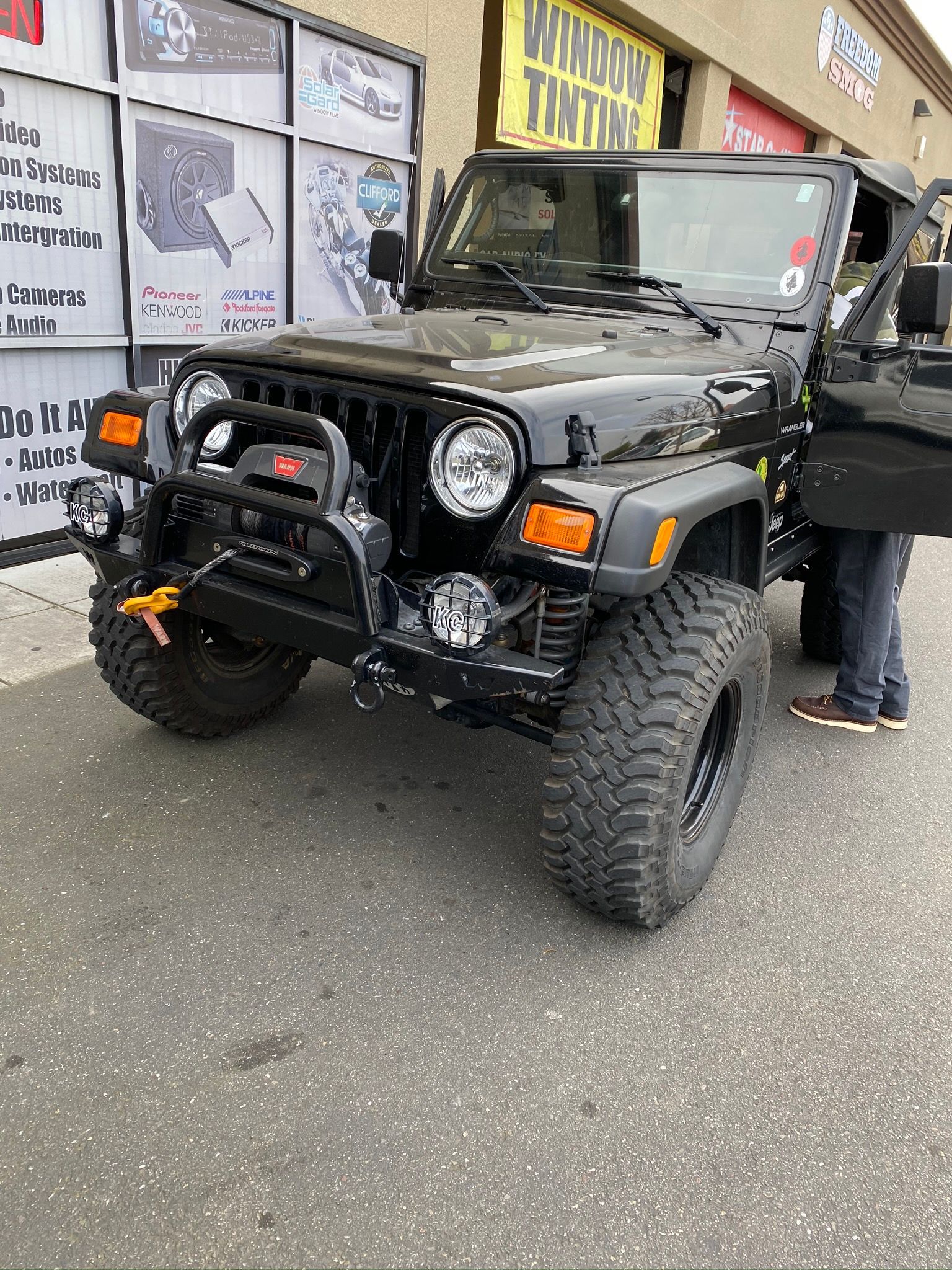 Black Jeep Wrangler with off-road tires parked outside a shop, front view with a person beside it