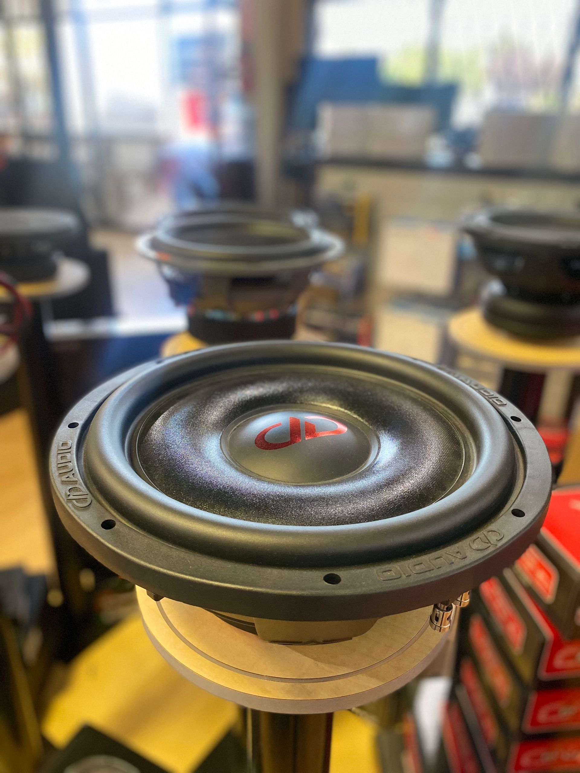 Close-up of a speaker driver with a red center cone on a workbench, blurred speakers in the background