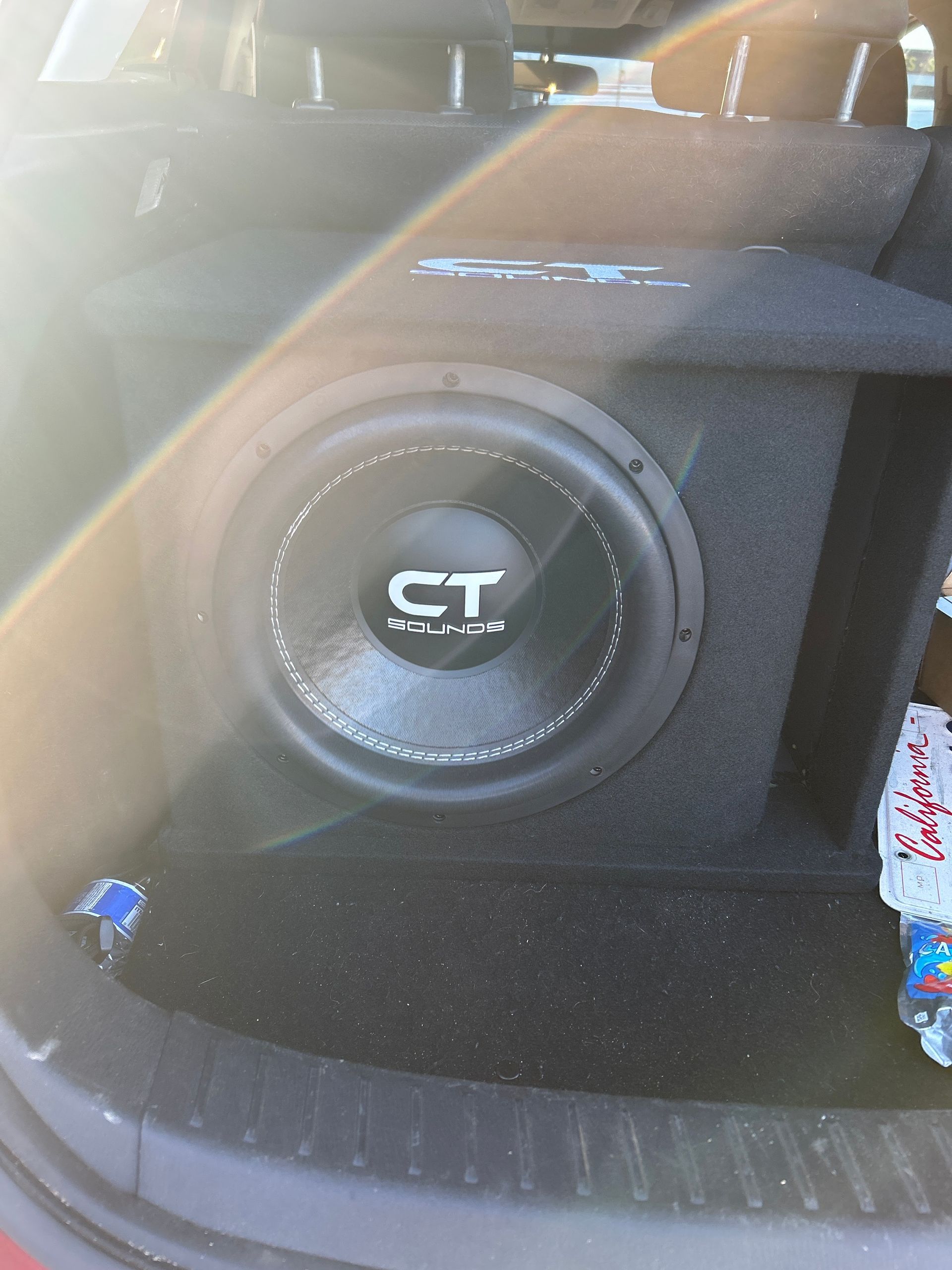 Car trunk with a large CT subwoofer speaker in a black enclosure, lit by sunlight