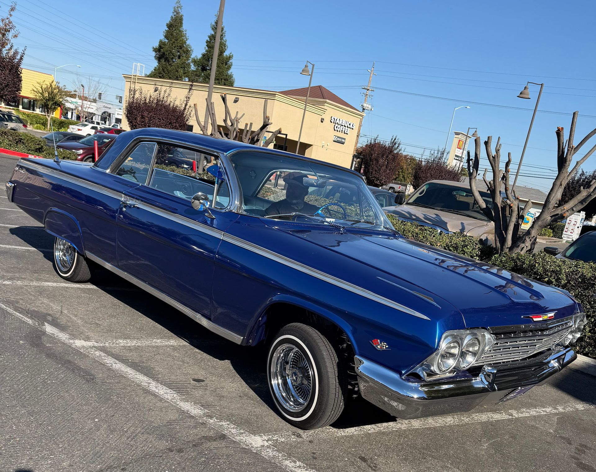 Blue classic car parked in a lot on a sunny day with buildings and trees in the background