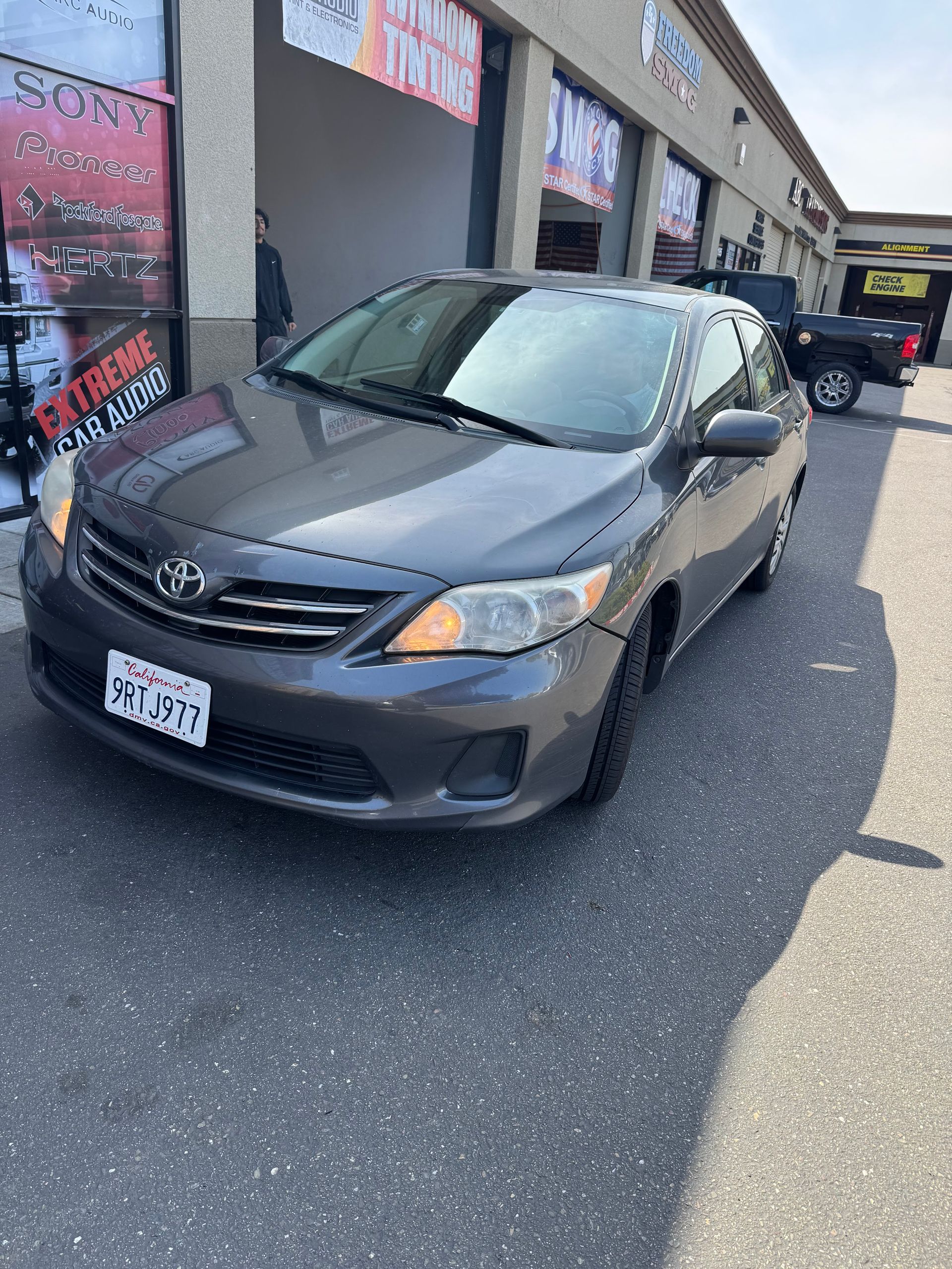 Gray Toyota sedan parked outside a strip mall on a sunny day, front view at an angle.