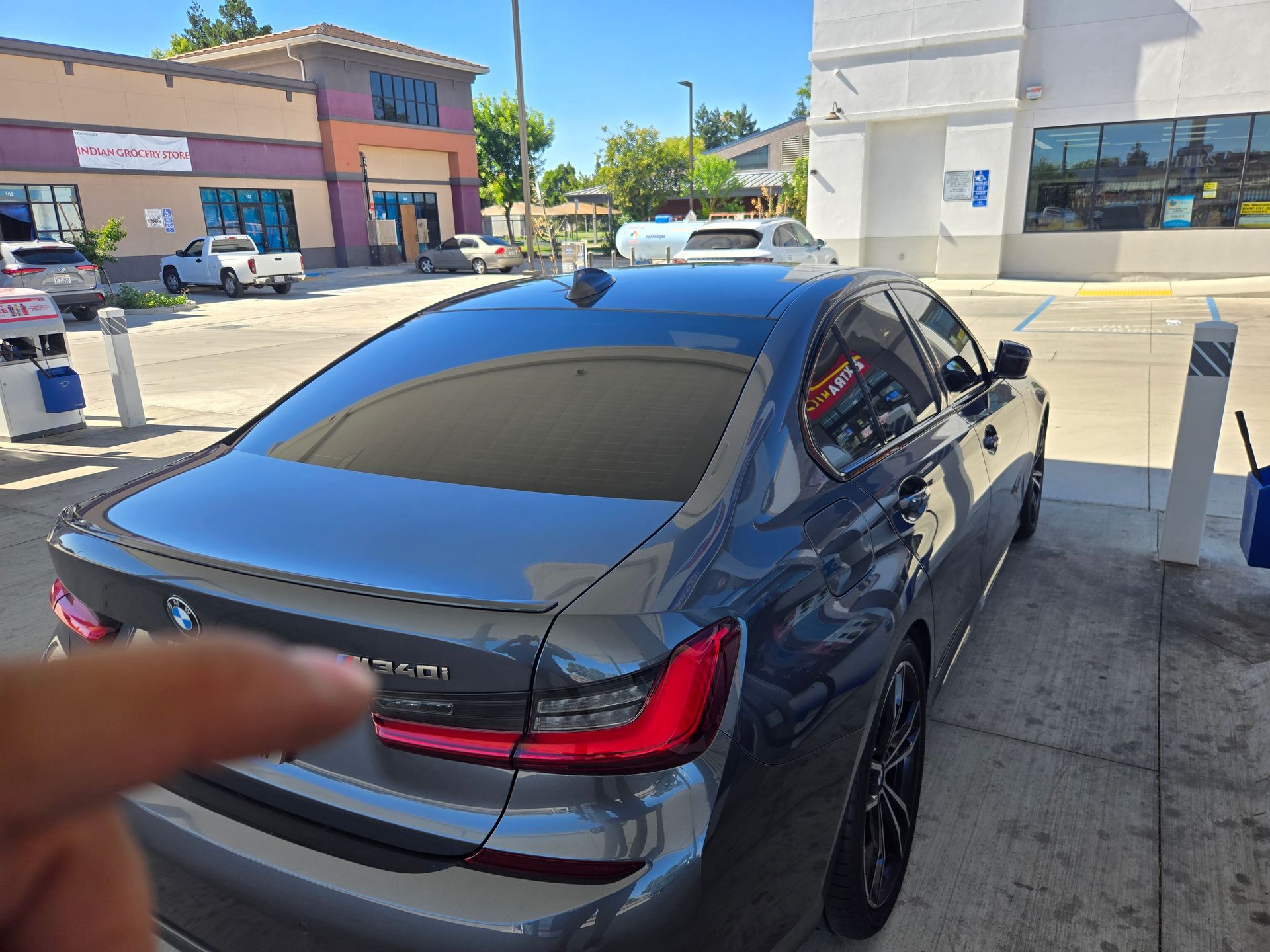 Dark gray sedan at a gas station, rear view with a finger pointing at the trunk badge.