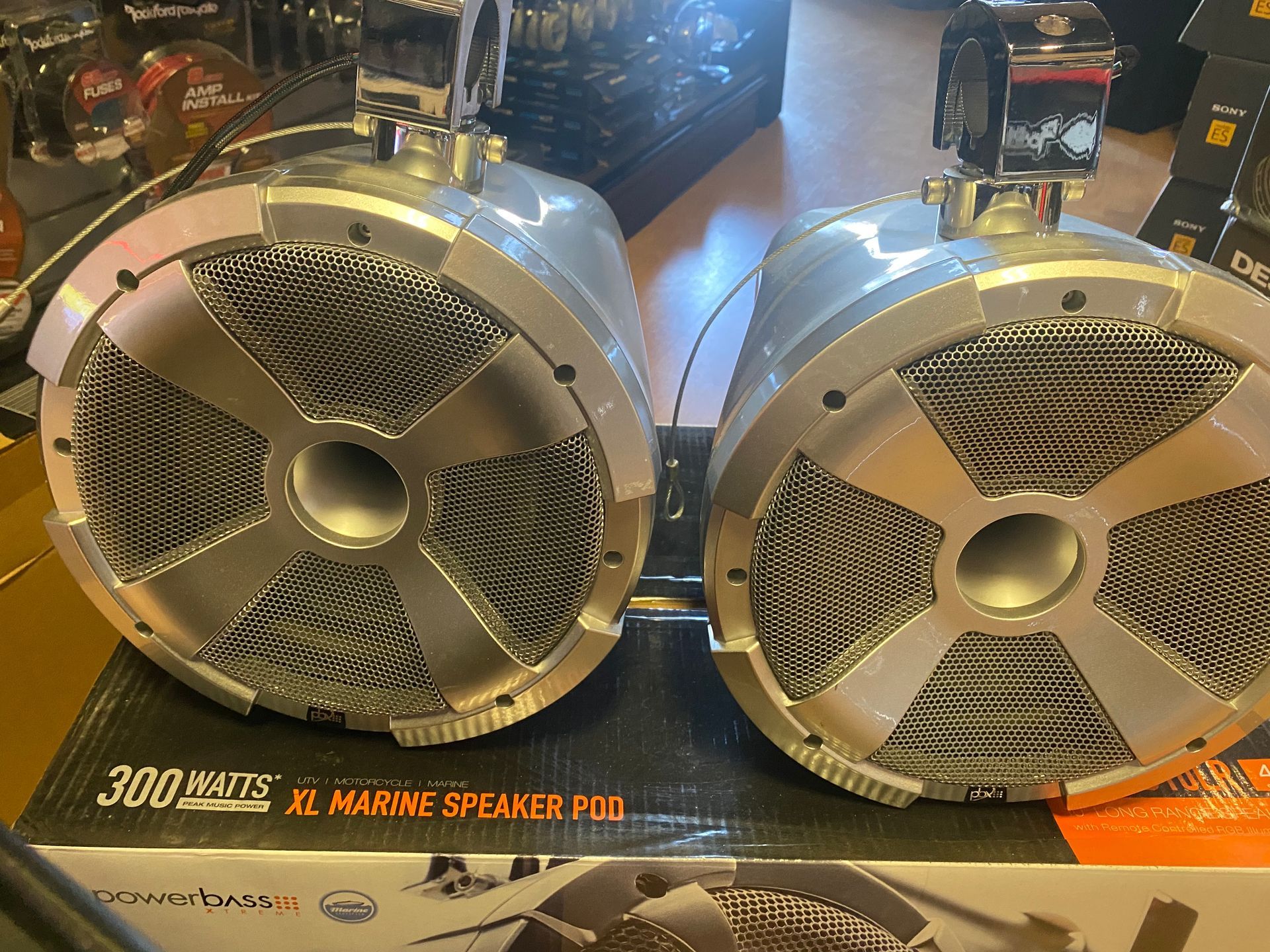 Two silver round speakers on a counter, with black mesh grilles and orange product packaging underneath