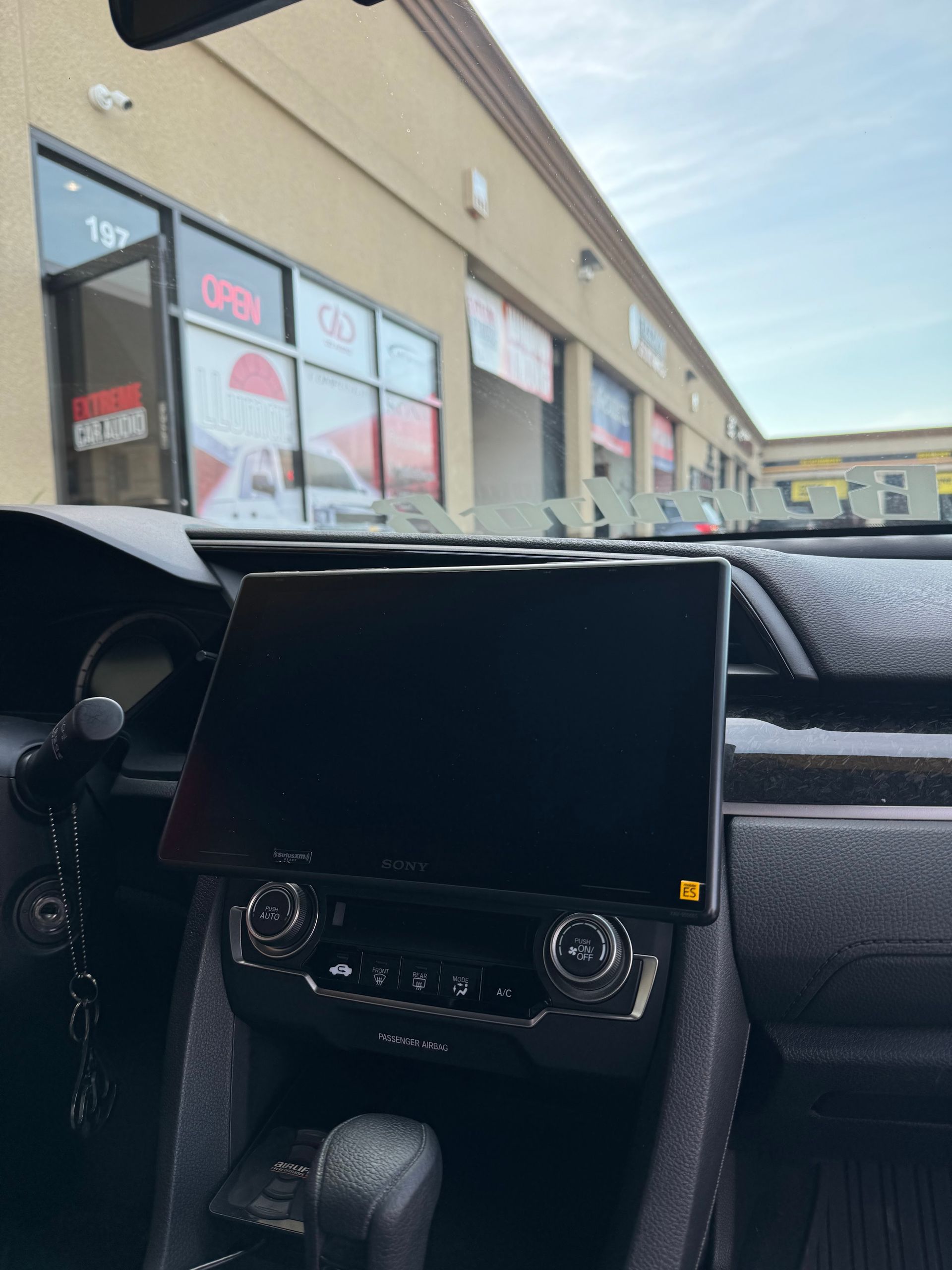 Car dashboard with a central touchscreen parked outside a strip mall storefronts.