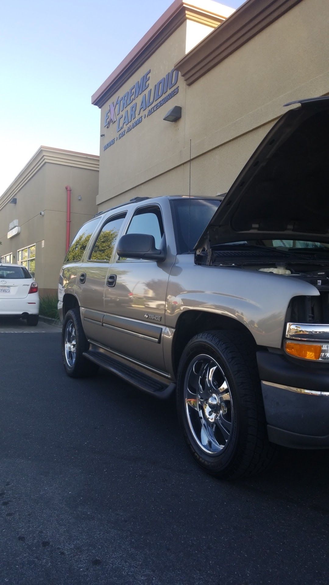 Silver SUV parked outside a beige building with its hood open, showing chrome wheels and a storefront sign above.