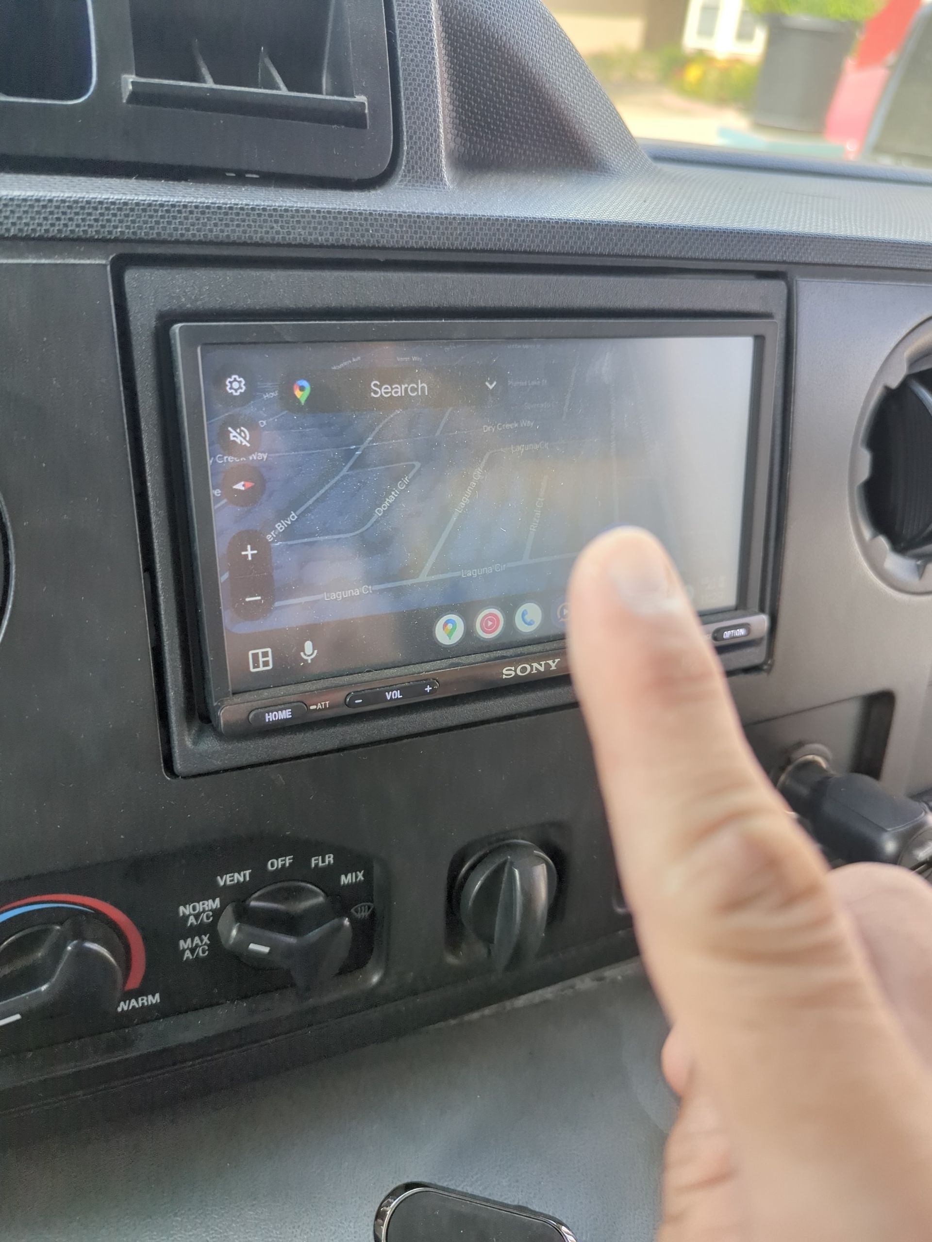 Finger pointing at a car touchscreen dashboard display with climate control knobs below.