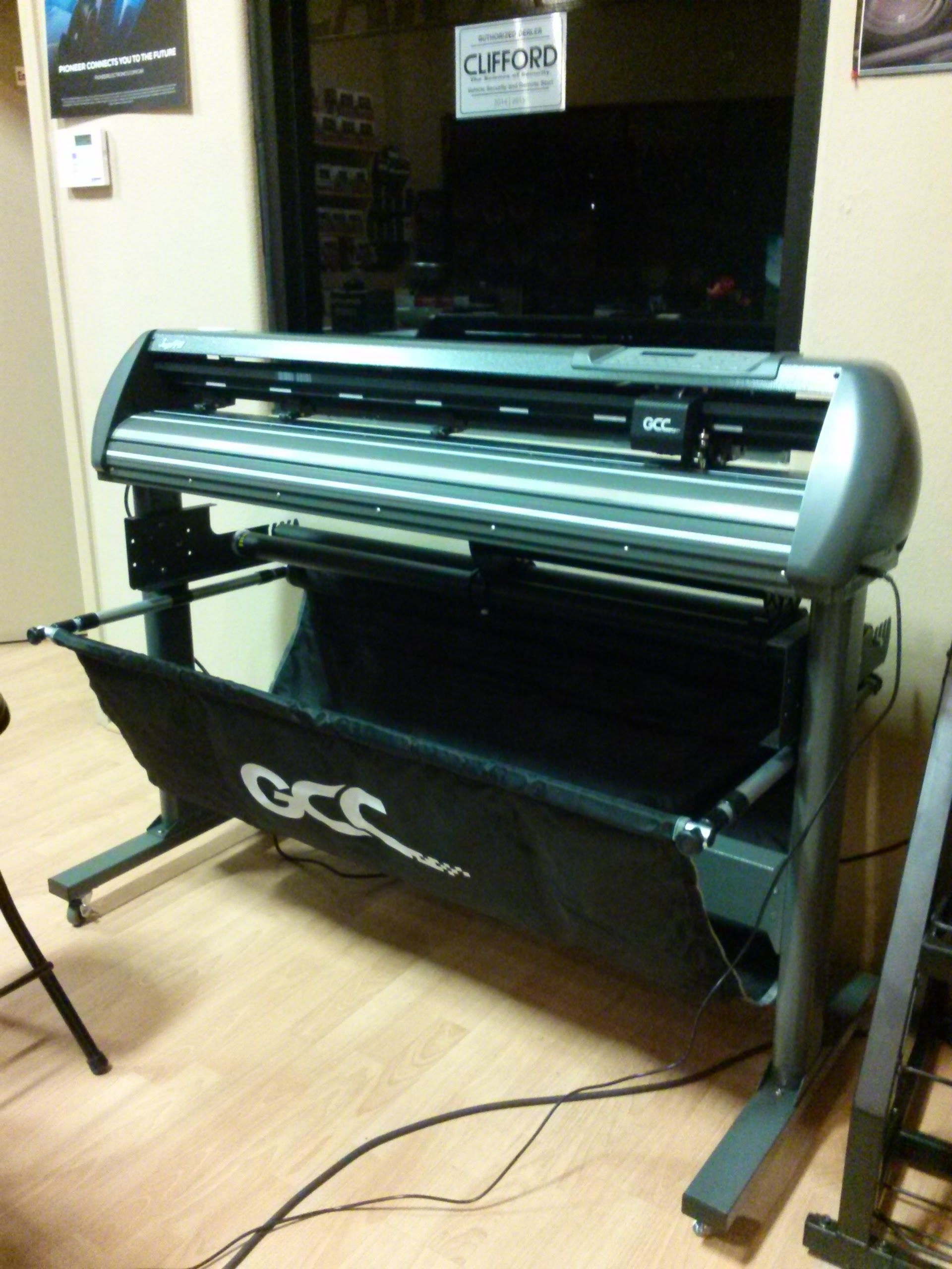 Large-format printer in a room, with a black banner hanging below it and cables on the floor.