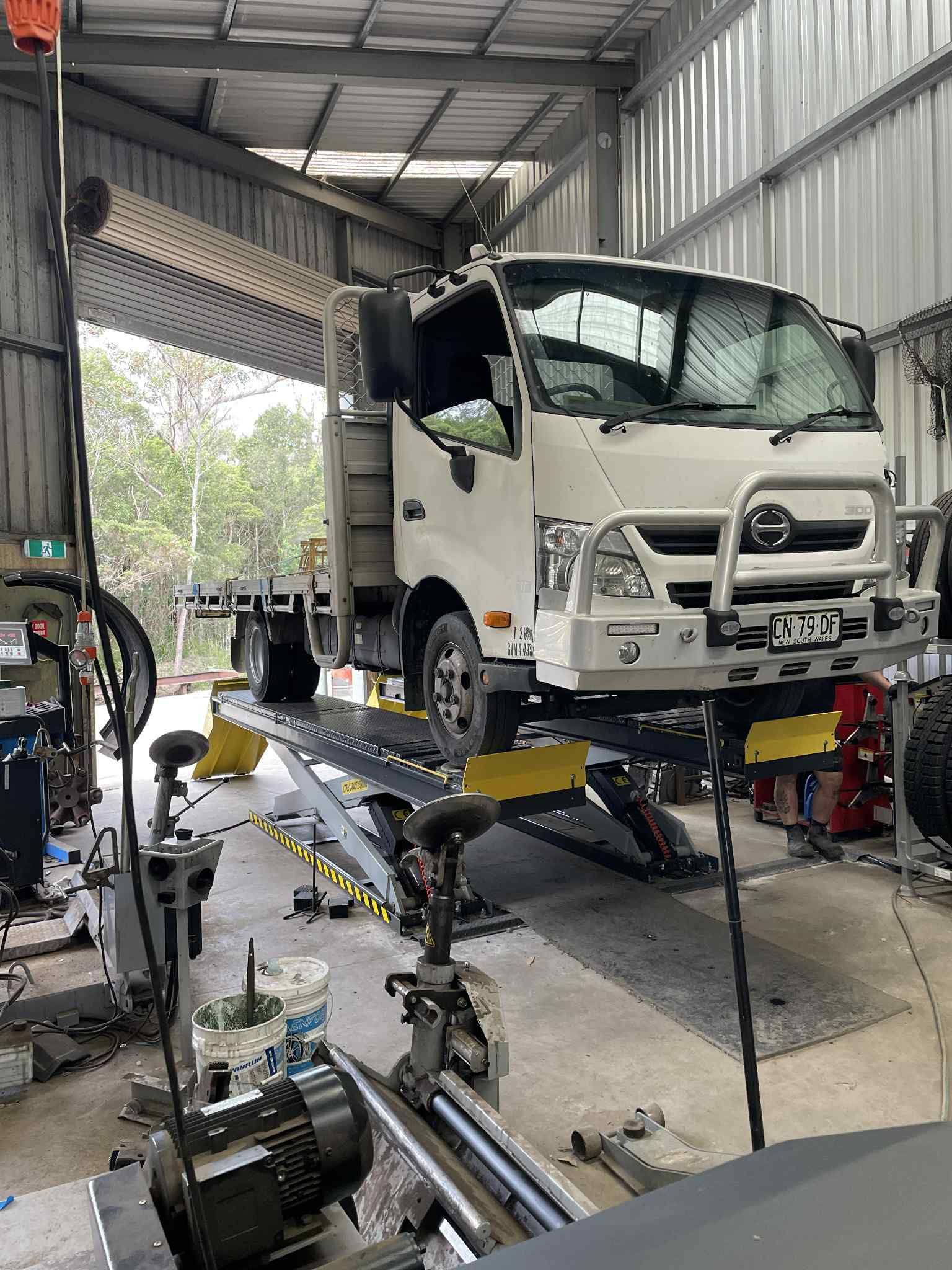 White truck raised on a repair lift inside an auto shop with tools and equipment nearby