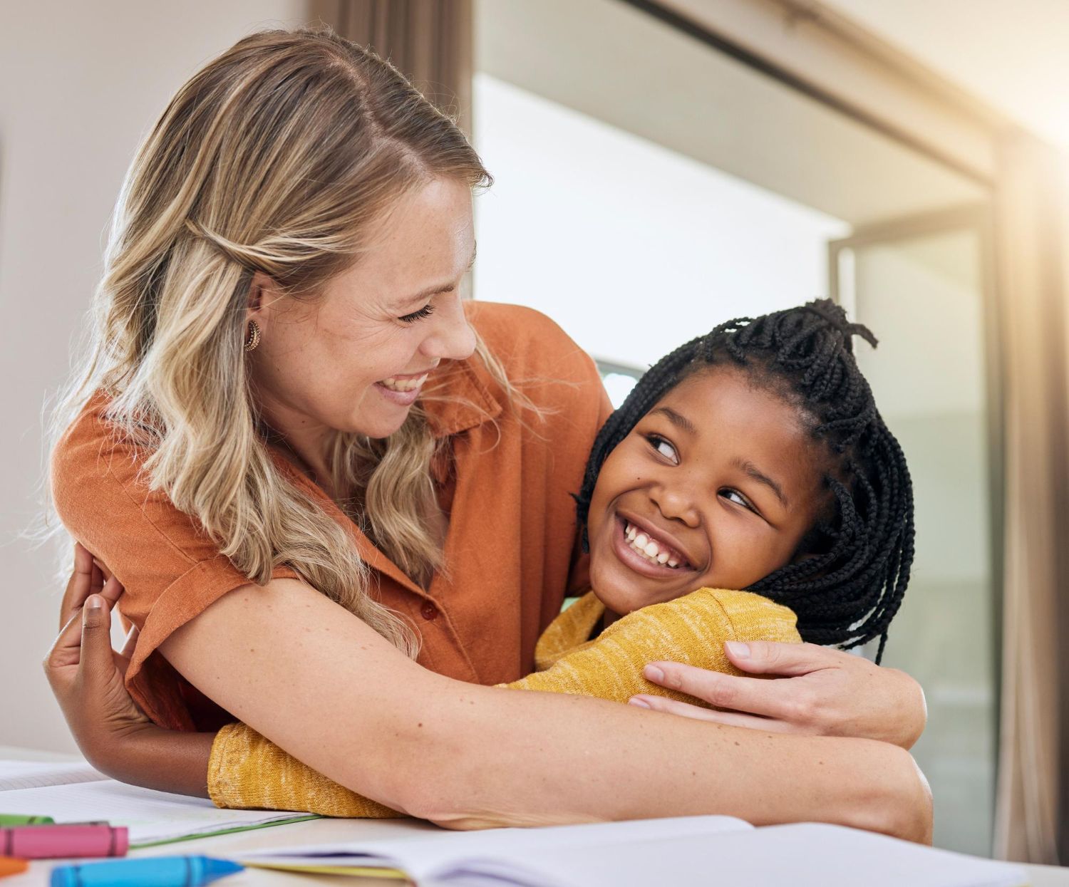 Woman hugging a smiling girl. They are both indoors, illuminated by sunlight.