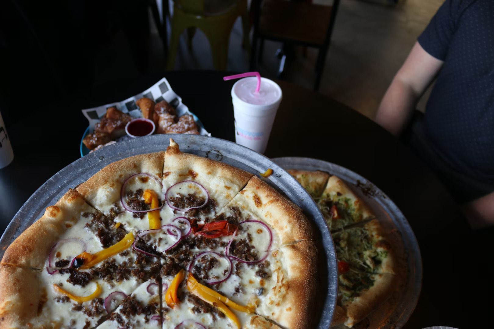 Two pizzas on metal trays, a basket of fried side dishes, and a drink with a pink straw on a dark table at a restaurant.
