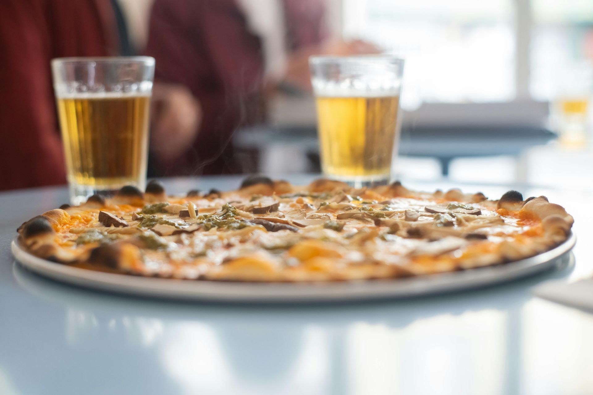 A mushroom pizza on a round white plate sits on a table in front of two glasses of beer.