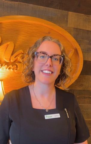 Woman with glasses smiles in front of a wood-textured wall and a gold-colored logo.
