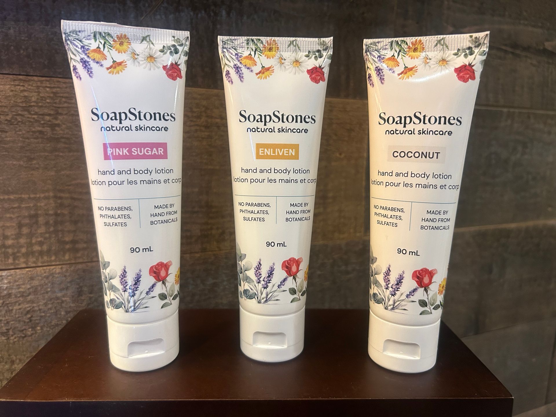 Three SoapStones body wash tubes in pink, yellow and white, on a wooden shelf.