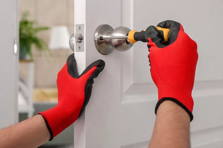 A Person Wearing Red Gloves Is Fixing a Door Handle with A Screwdriver — Coffs City Lockmart Pty Ltd in North Boambee Valley, NSW