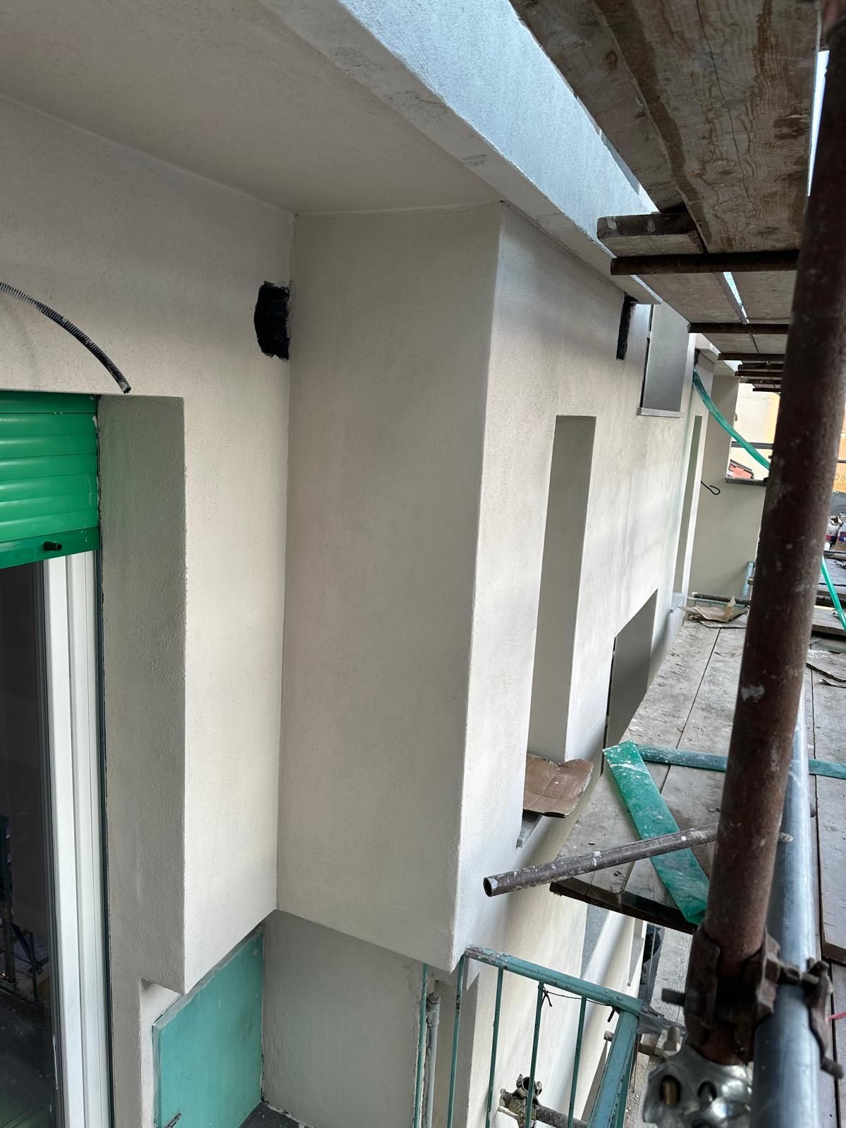 Stretto corridoio esterno con pareti bianche, porta verde e impalcature sul lato destro.