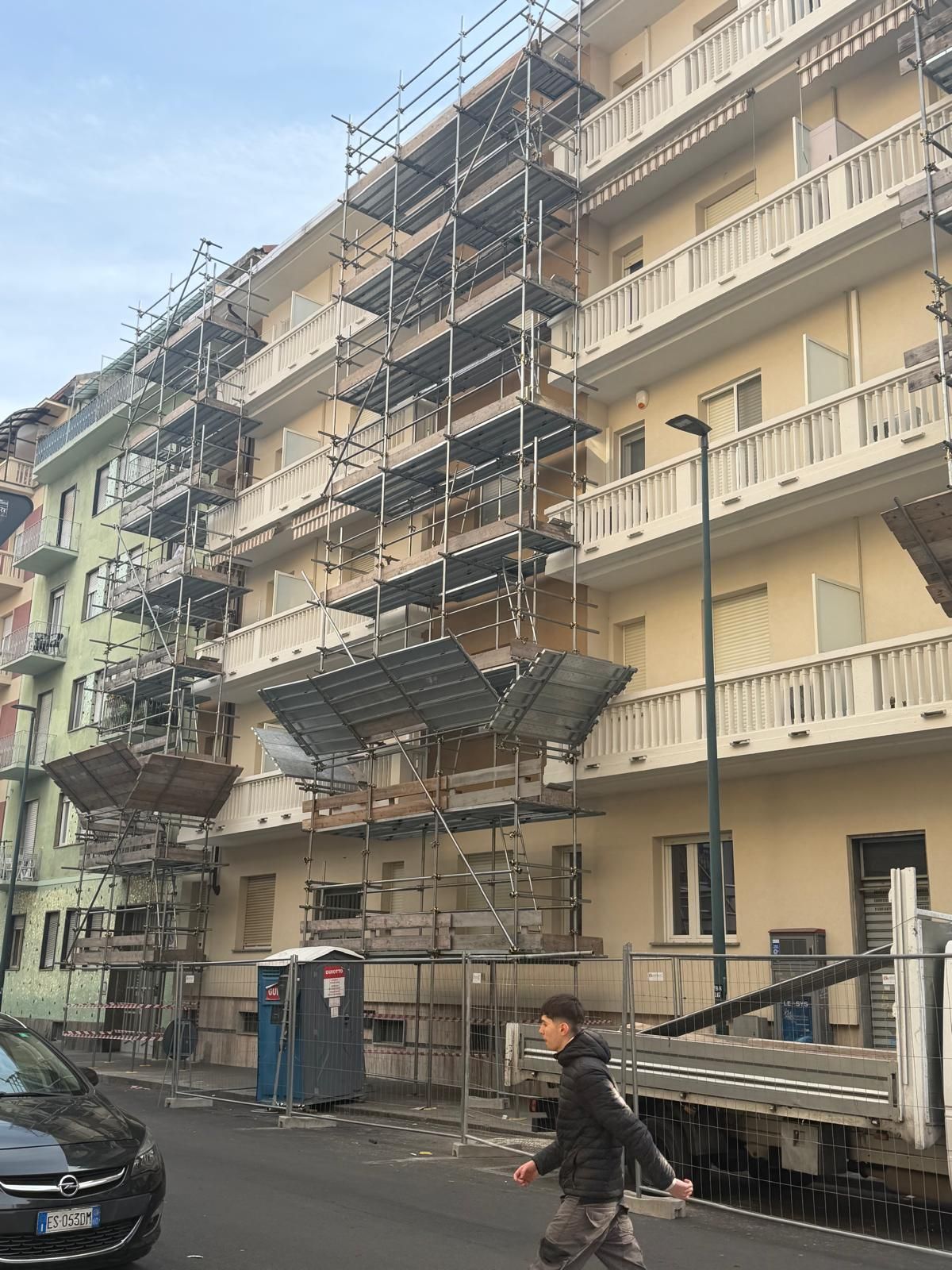 Ponteggi sulla facciata di un condominio con piattaforma per gli operai e un pedone sulla strada sottostante.