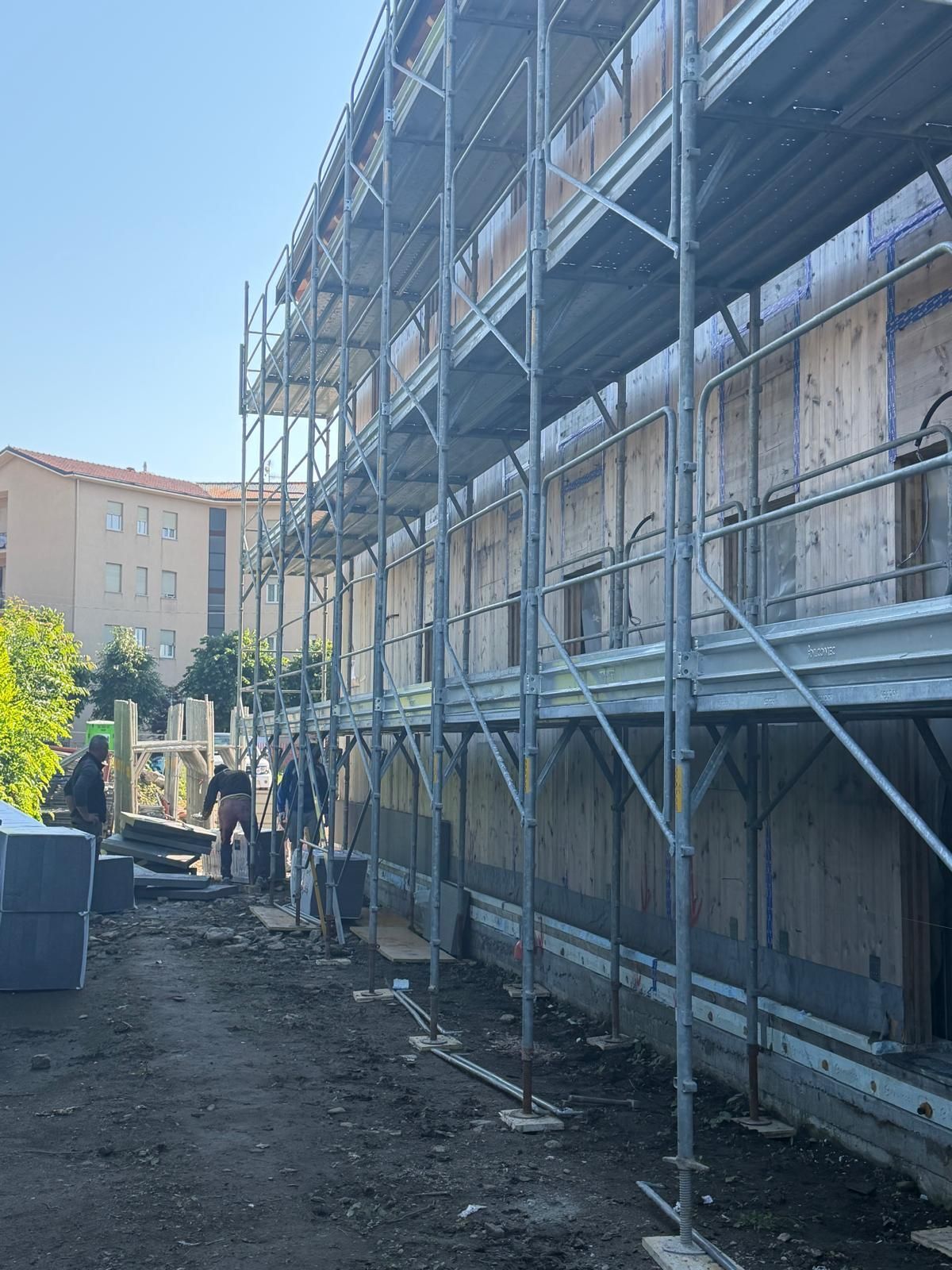 Impalcature lungo la facciata di un edificio, accanto a un sentiero sterrato e in lontananza a dei palazzi residenziali, in una giornata di sole.