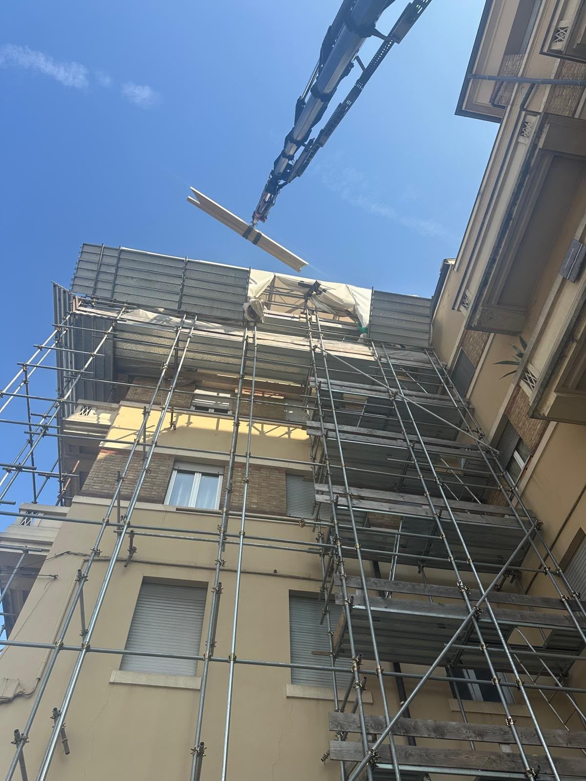 Impalcature che ricoprono un edificio color sabbia sotto un cielo azzurro, con il braccio di una gru sopra la testa.