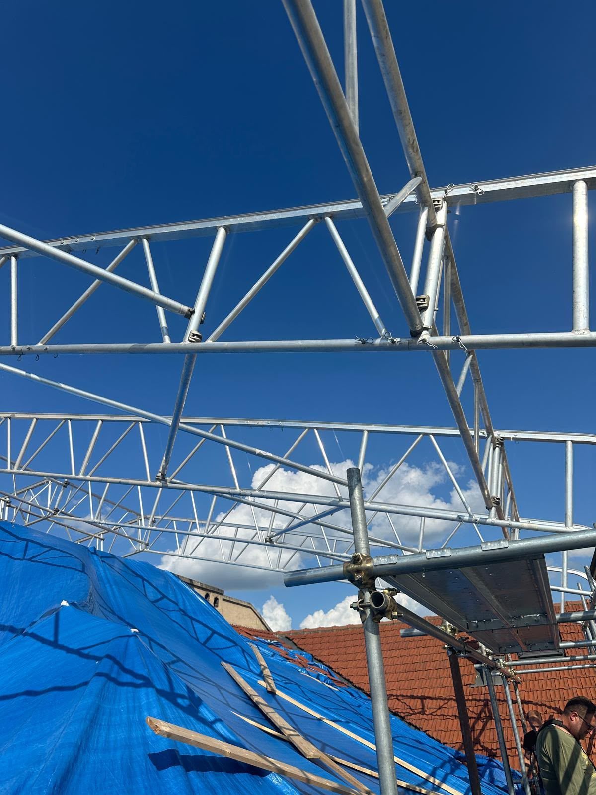 Tetto a capriate metalliche parzialmente assemblato sopra un telo blu e edifici in mattoni sotto un cielo azzurro limpido.