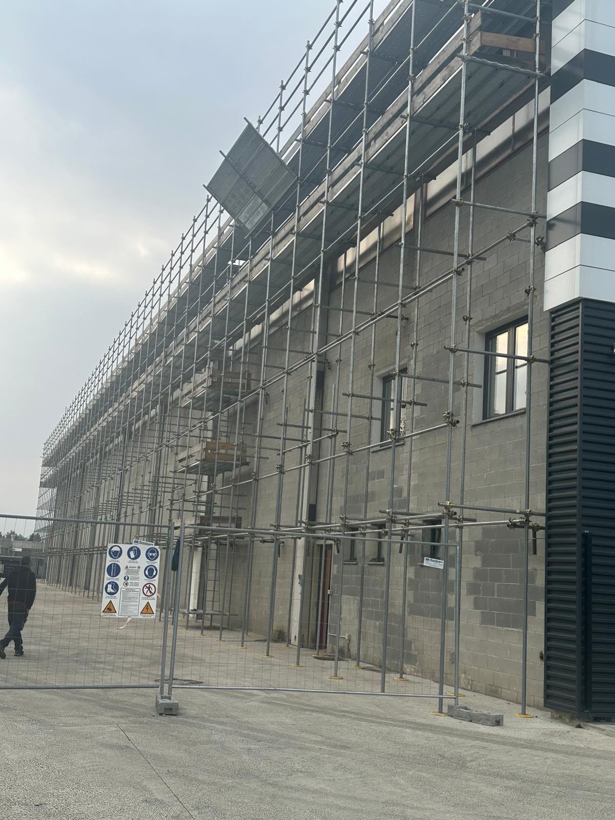 Edificio in costruzione con impalcature lungo la facciata e un cartello pedonale di fronte.