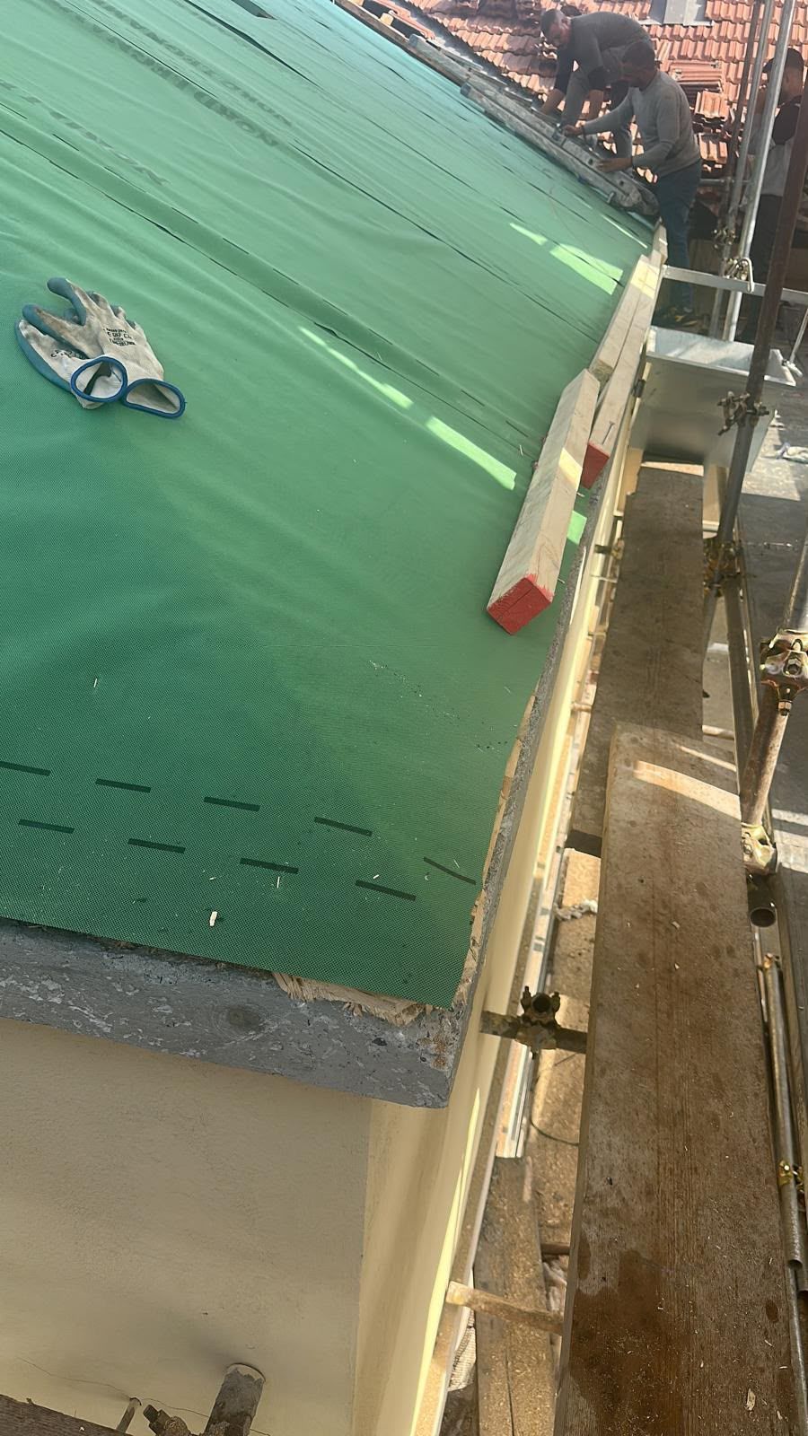 Vista inclinata di un tetto coperto da un telone verde, con un piccolo ventilatore e una scala lungo il bordo, in un cantiere edile.