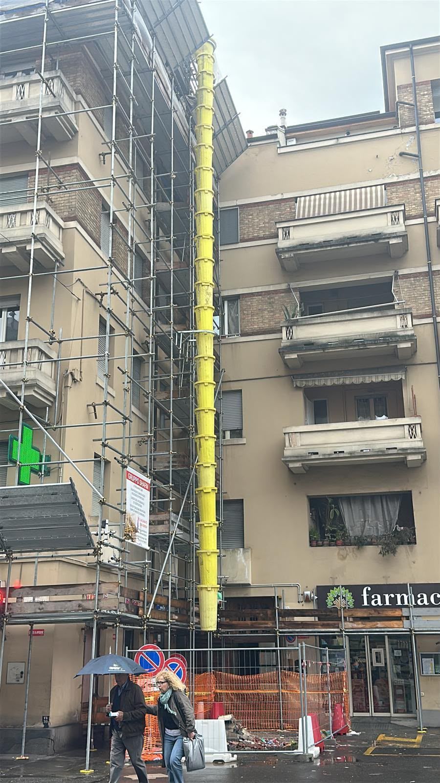 Scena di strada con un edificio ricoperto di impalcature, un tubo verticale giallo e alcuni pedoni vicino a una vetrina.