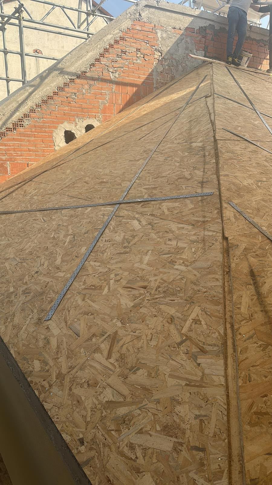 Rivestimento del tetto in legno in fase di costruzione, con linee di supporto blu e un muro di mattoni sullo sfondo.