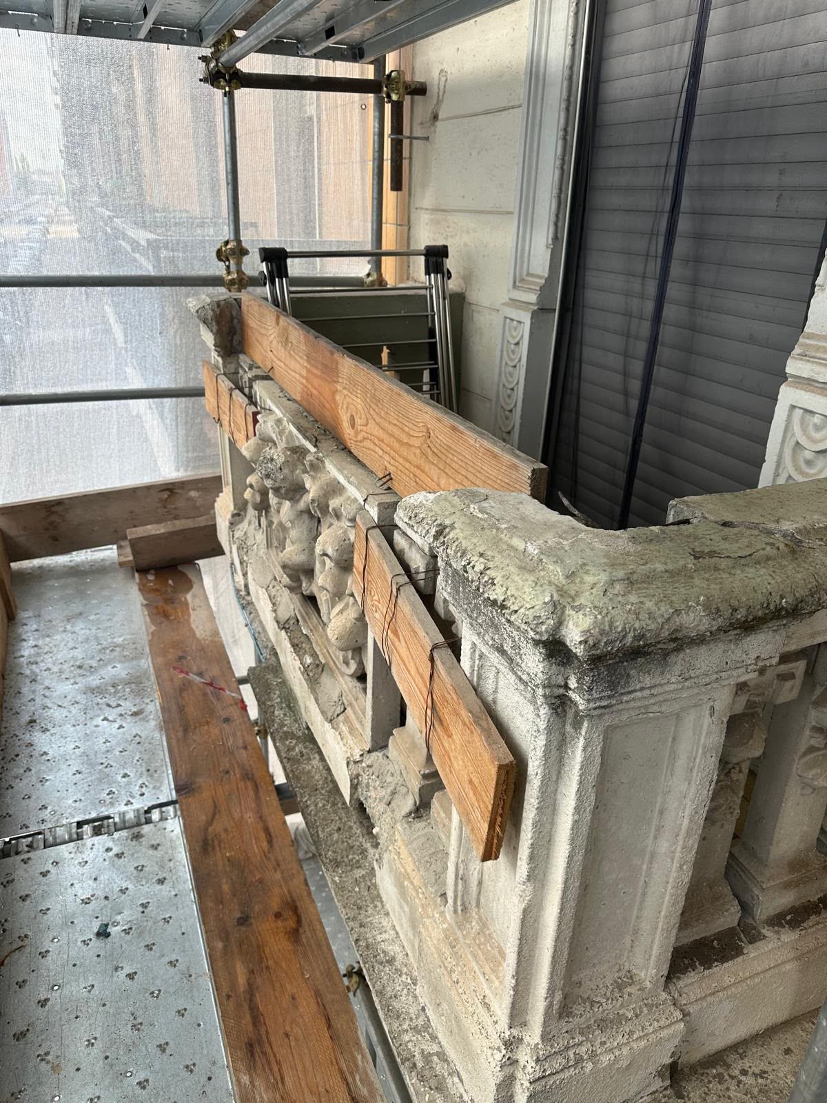Impalcature accanto a un muro di pietra grezza con assi di legno e un telo grigio su un pavimento bagnato.