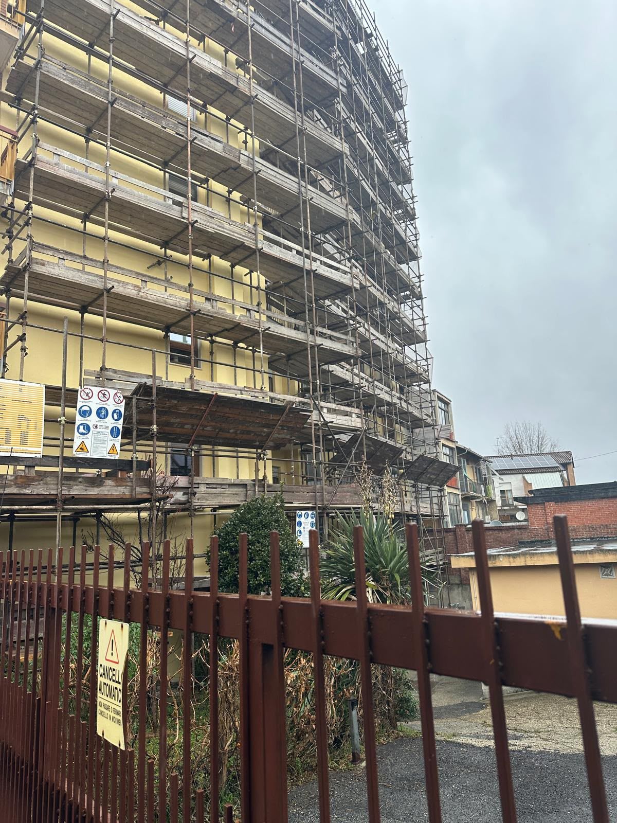 Palazzo di appartamenti curvo in fase di ristrutturazione, con impalcature accanto a una recinzione e cielo nuvoloso.