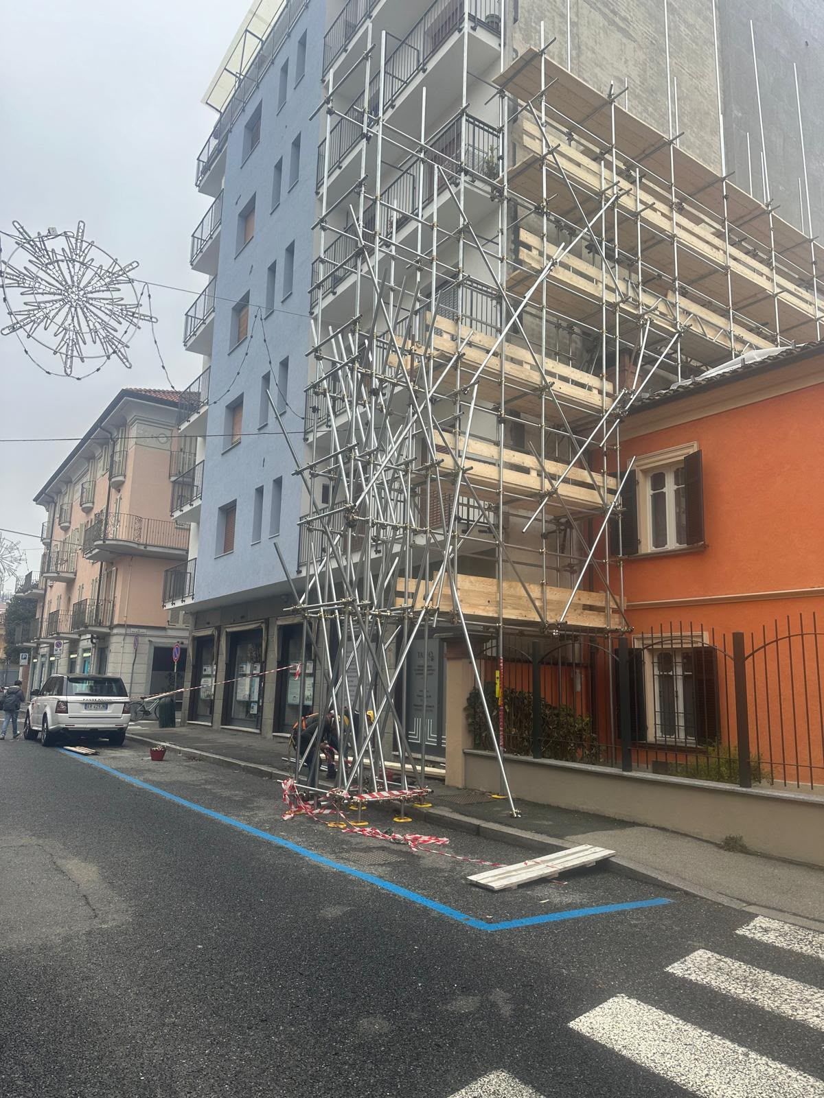 Angolo di strada con un edificio blu sotto impalcature e un'auto bianca parcheggiata nelle vicinanze.