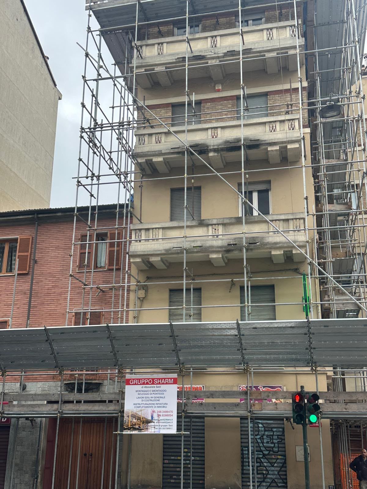 Un condominio beige, con impalcature, è in fase di ristrutturazione accanto a un edificio in mattoni, con una vetrina e un semaforo sottostanti.