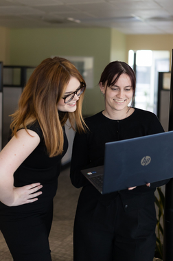 Two people in black clothing stand in an office, looking at a laptop screen together and smiling.