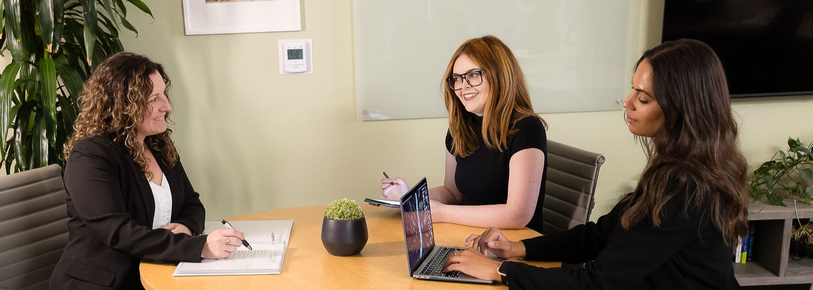 Three people sit at a round table in an office, working on documents and a laptop during a professional meeting.