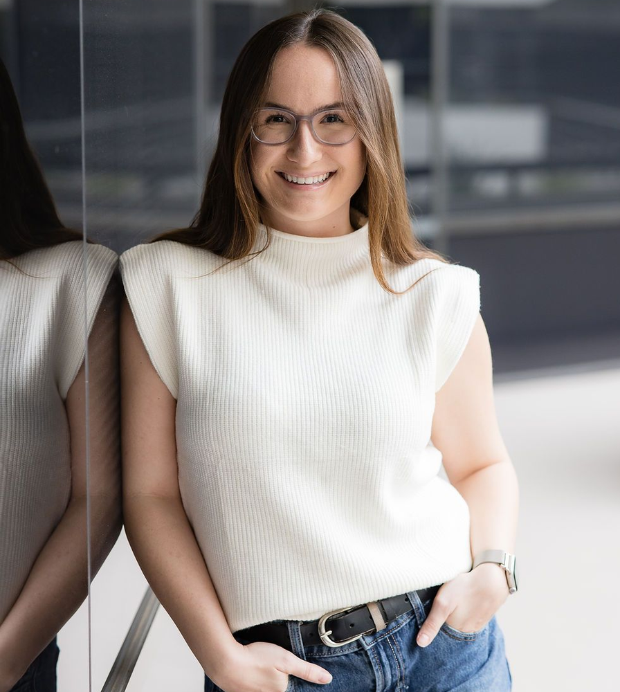 A smiling person with long hair and glasses wears a white sweater and jeans, posing next to a dark glass wall.