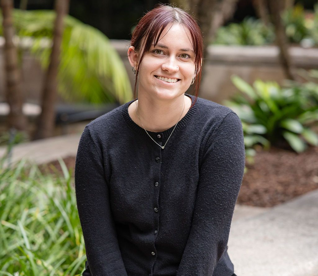 A smiling person with reddish hair wearing a dark cardigan, sitting outdoors in a garden setting.