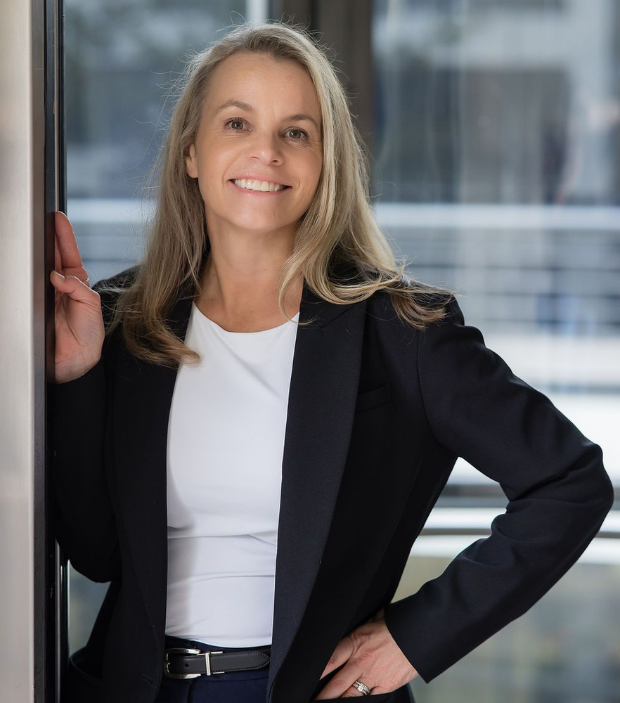 Smiling woman wearing a black blazer over a white shirt, leaning against a door frame in a bright, modern office.