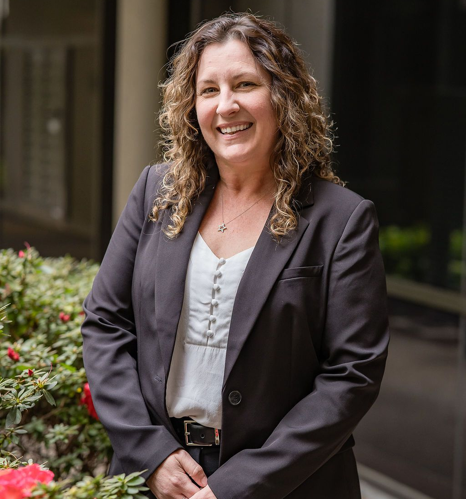 A smiling person with curly hair wears a dark blazer and white blouse, standing outdoors near a flowering bush.
